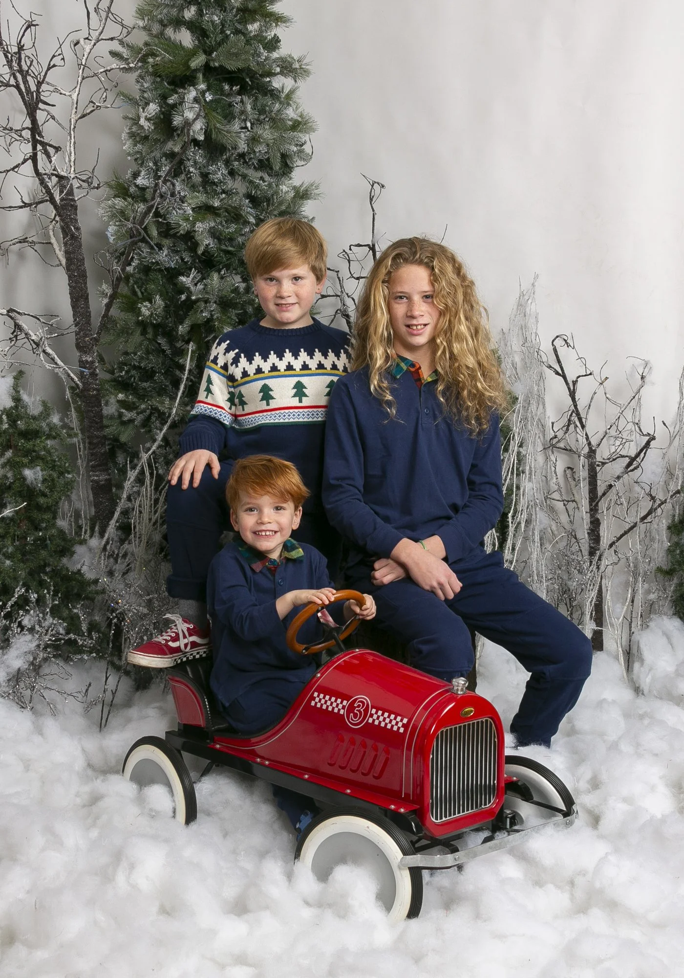 Three siblings in festive outfits sitting in a vintage red pedal car on a winter wonderland set at Hayes & Fisk’s portrait studio in Richmond.
