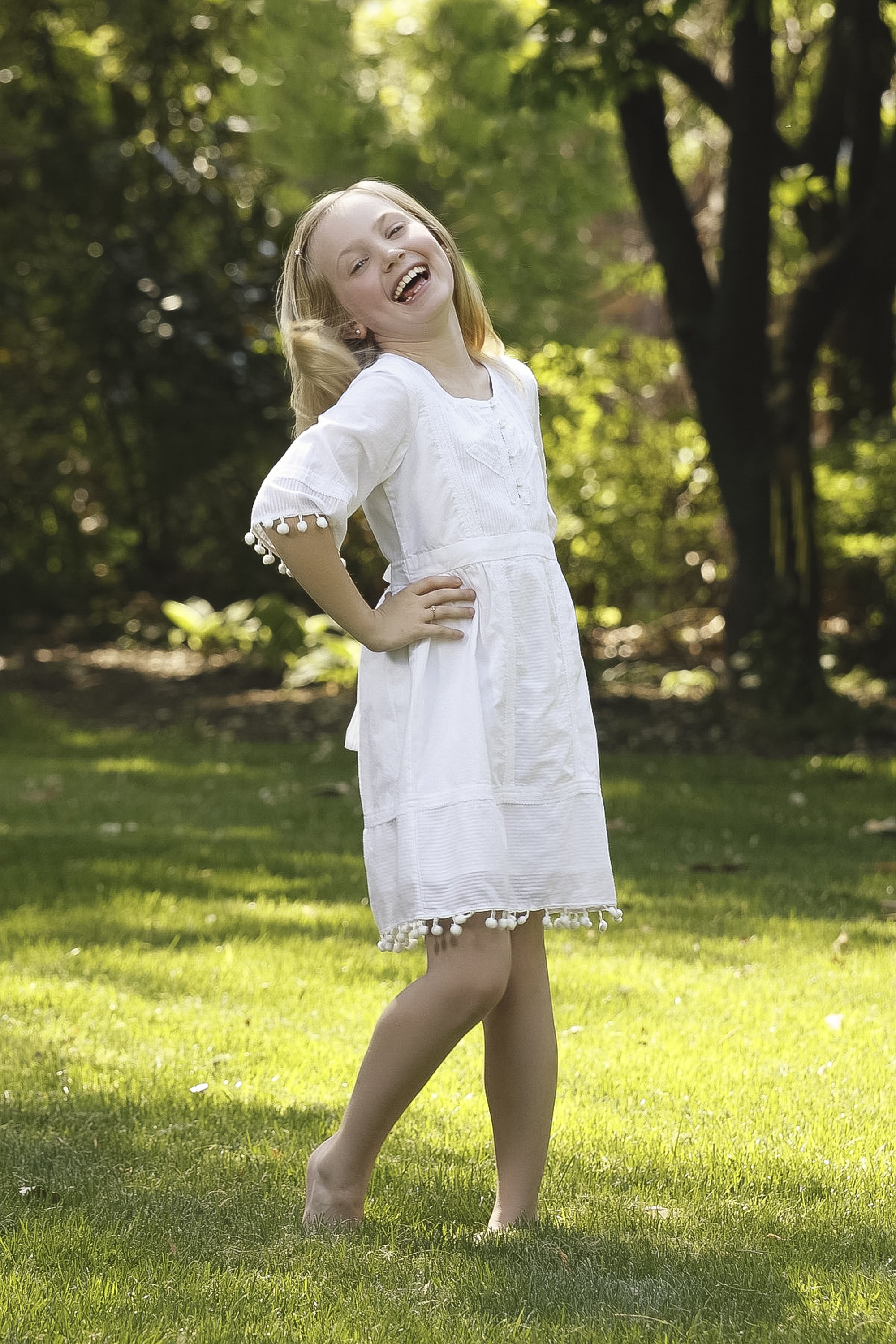 Young blonde girl laughing with her hands on her hips, wearing a white dress while standing outdoors in a sunlit garden.
