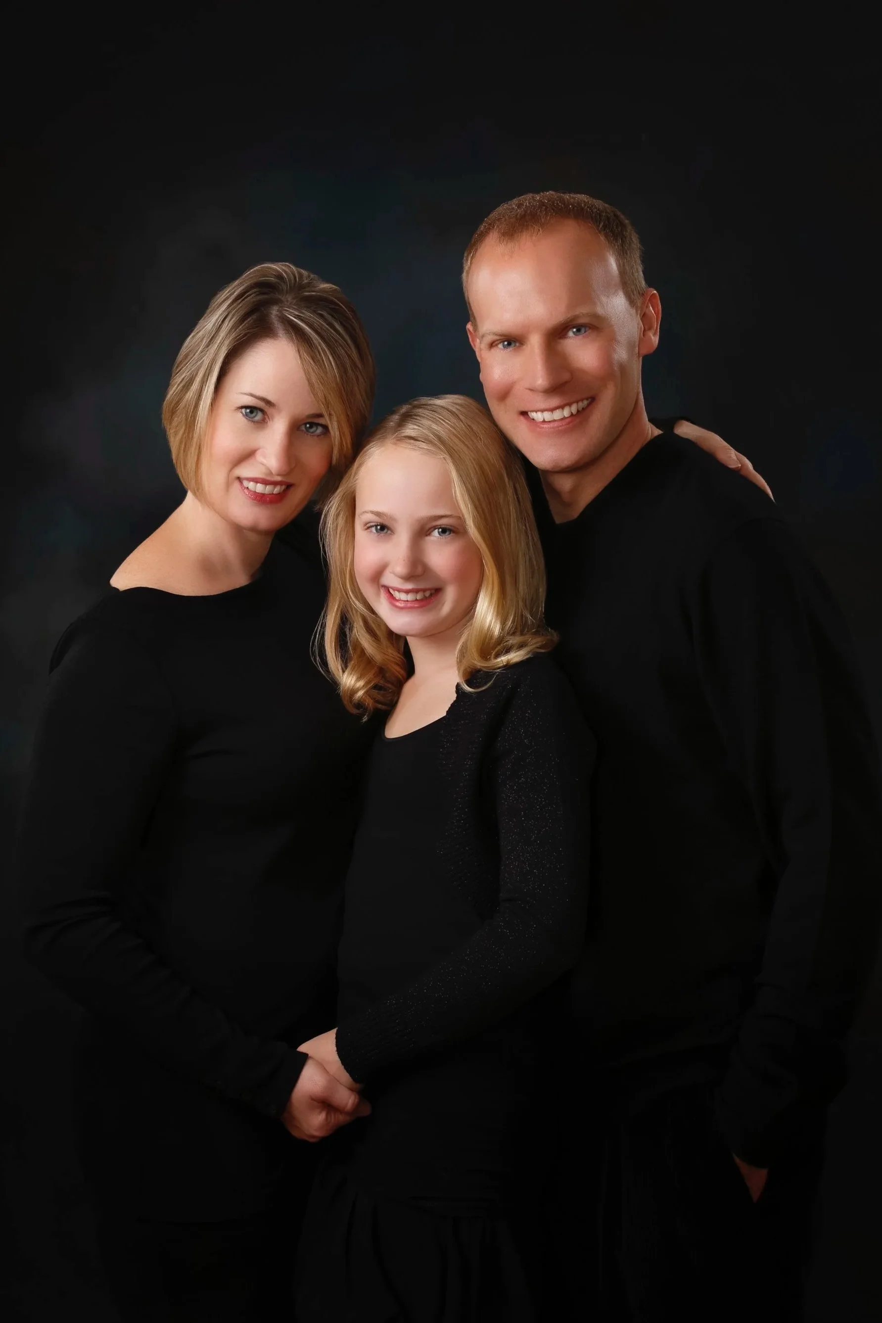 Studio portrait of a mother and father with their blonde daughter, all dressed in black against a dark background.
