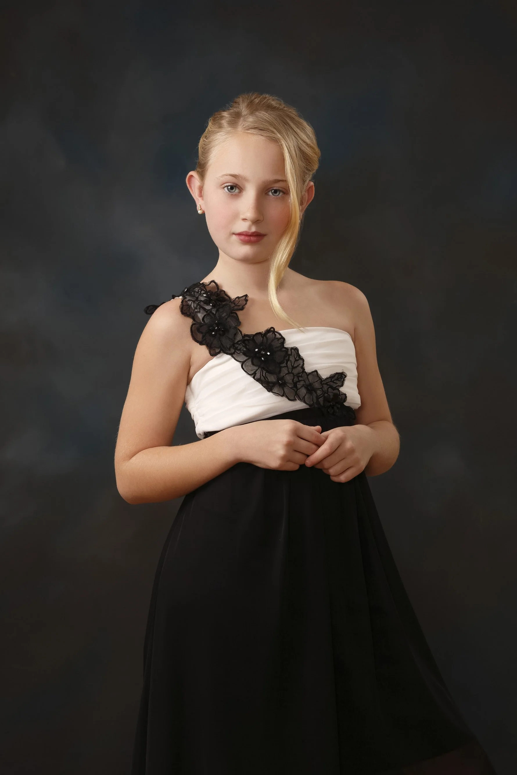 Young blonde girl wearing a formal black-and-white dress with black floral detailing, posed against a dark studio background.