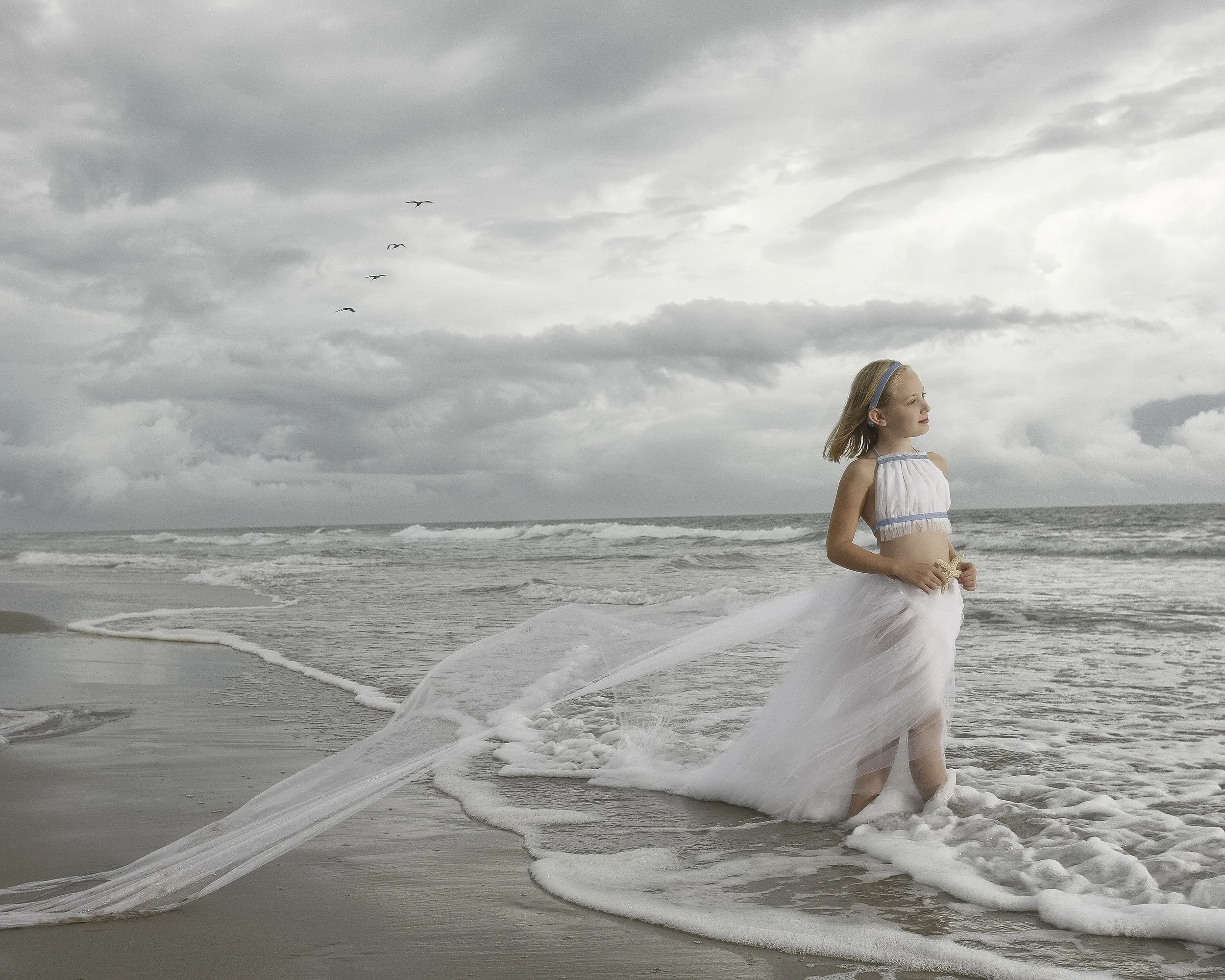 Blonde girl standing at the ocean's edge in a pale blue and white outfit with flowing tulle, beneath a cloudy sky.
