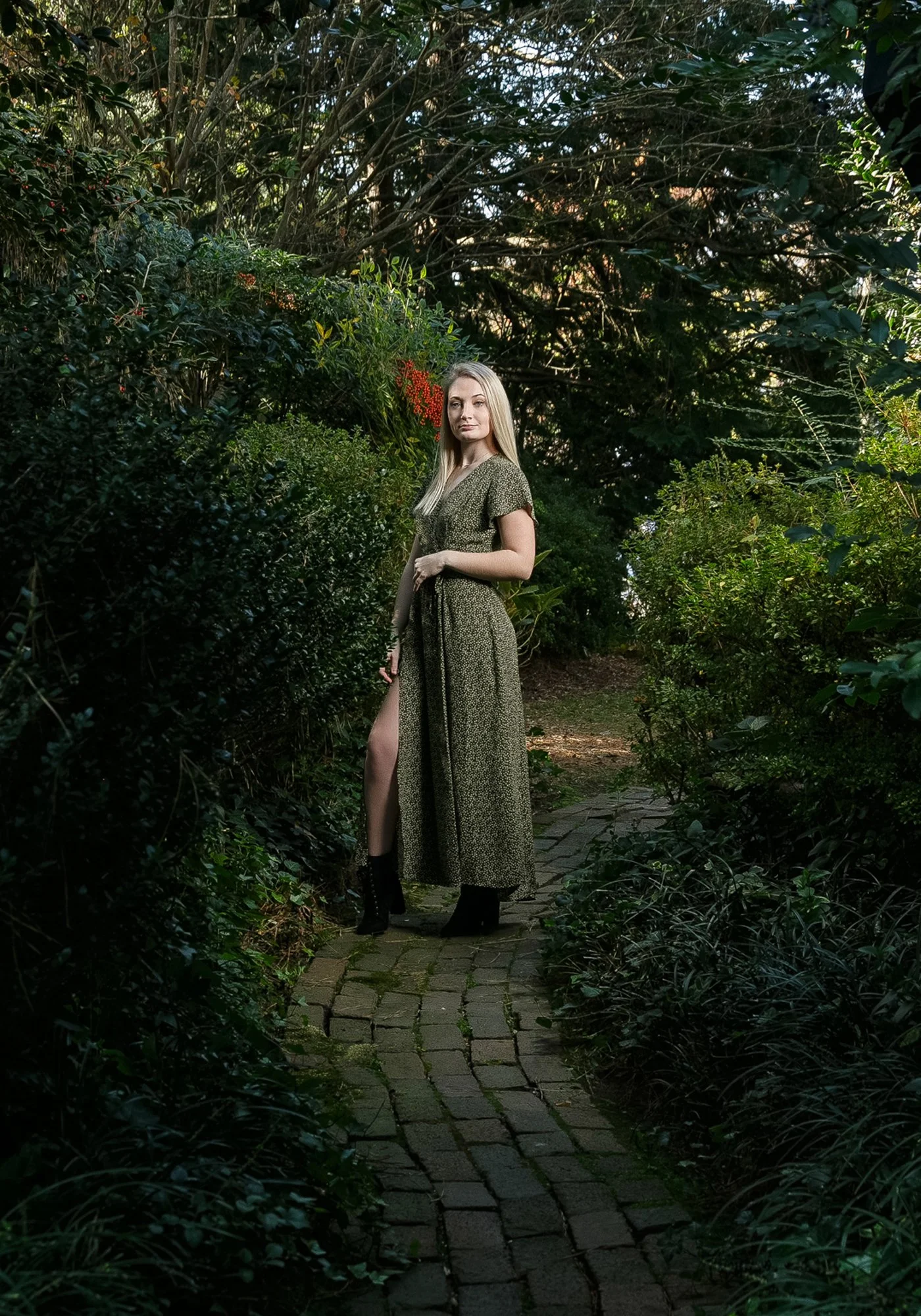 Young blonde woman wearing an olive-green dress and black boots, standing on a shaded stone garden path surrounded by greenery.