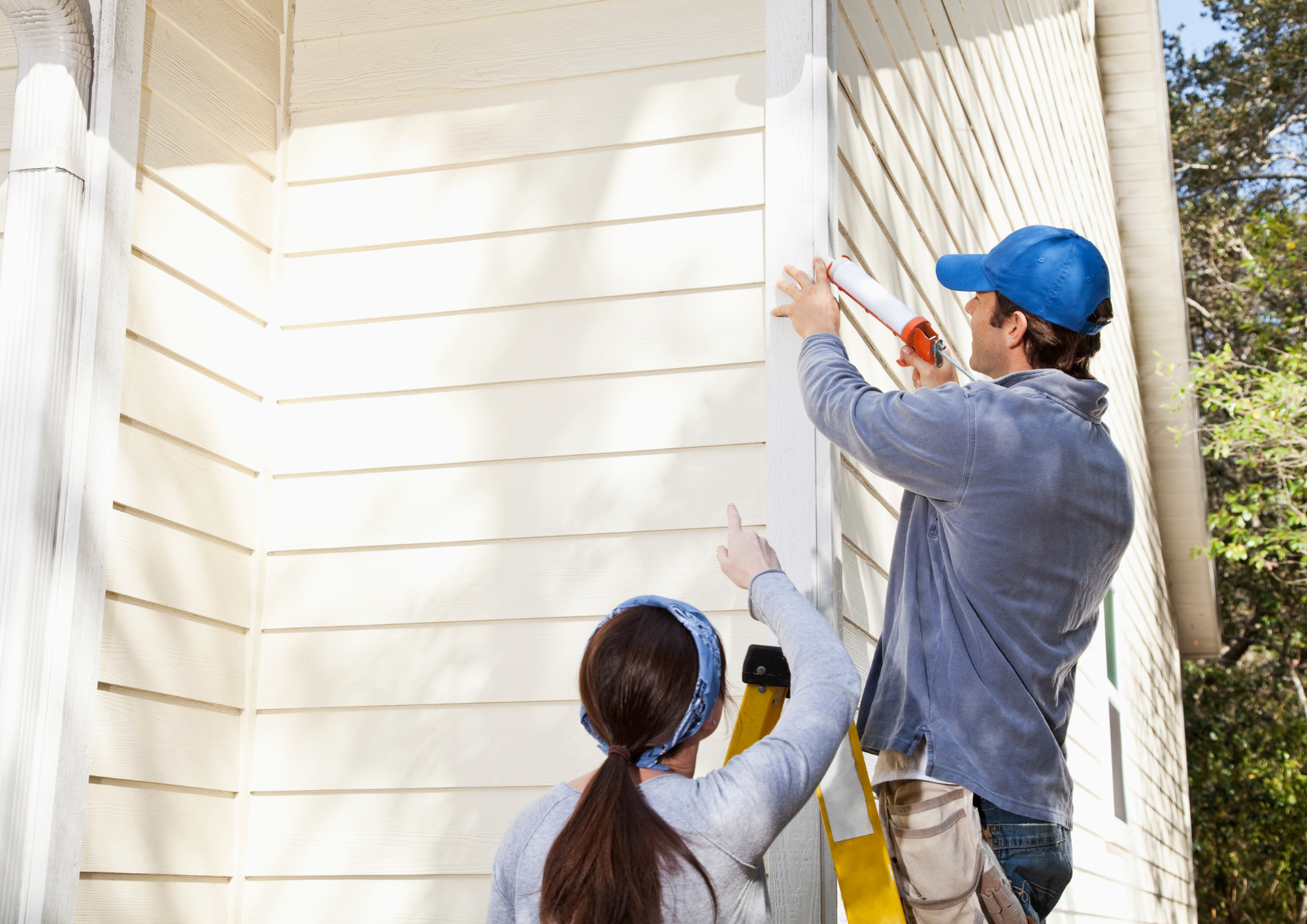 A man is applying caulking to the exterior of a house with siding. A woman with long brown hair wearing a bandana is standing below & pointing up, while another person in the background holds a yellow ladder. NDIS Home Maintenance Adelaide.
