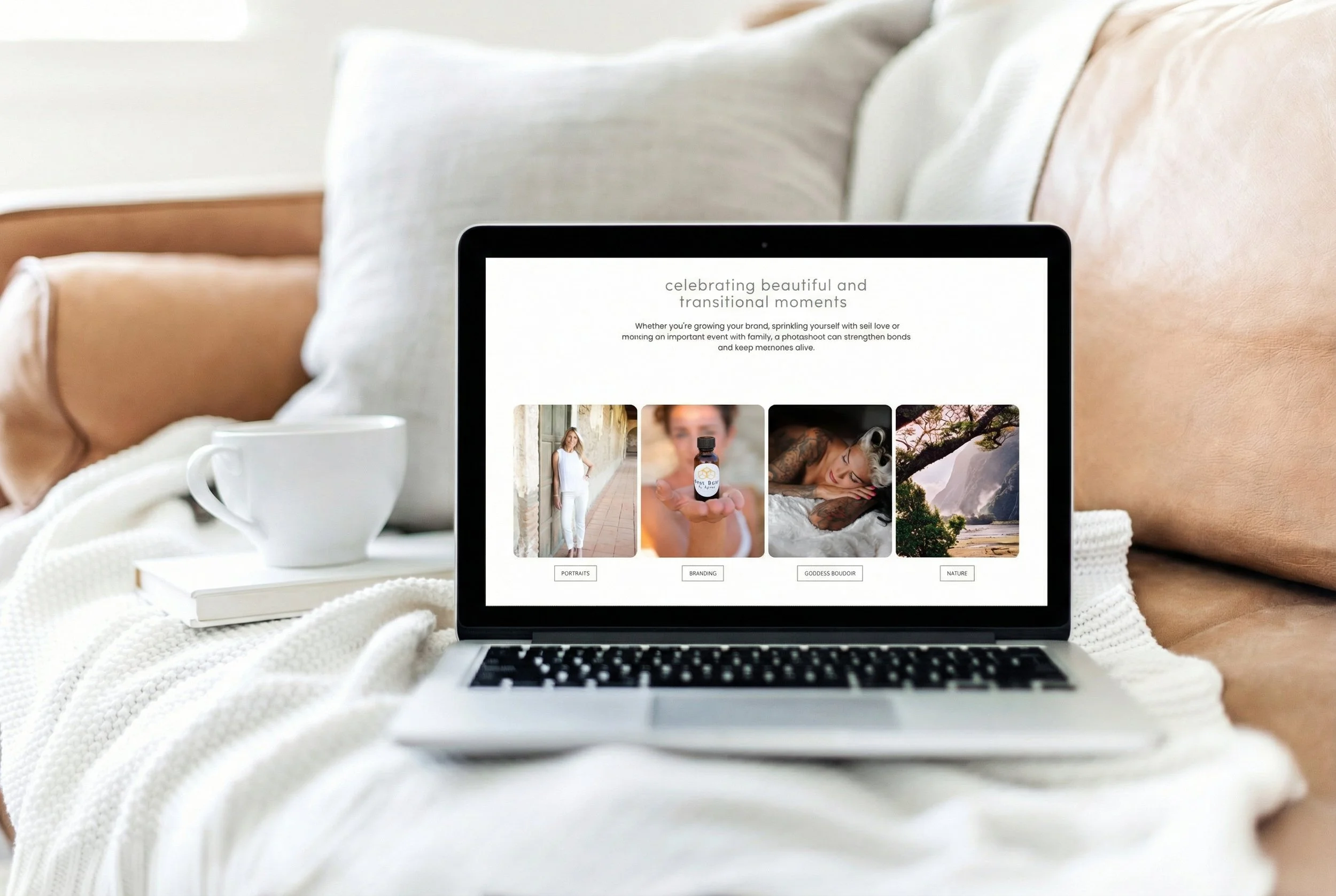 Laptop displaying a website about celebrating beautiful and transitional moments, with categories portraits, branding, goddess boudoir, and nature, placed on a cozy white blanket on a couch, next to a white cup and a book.