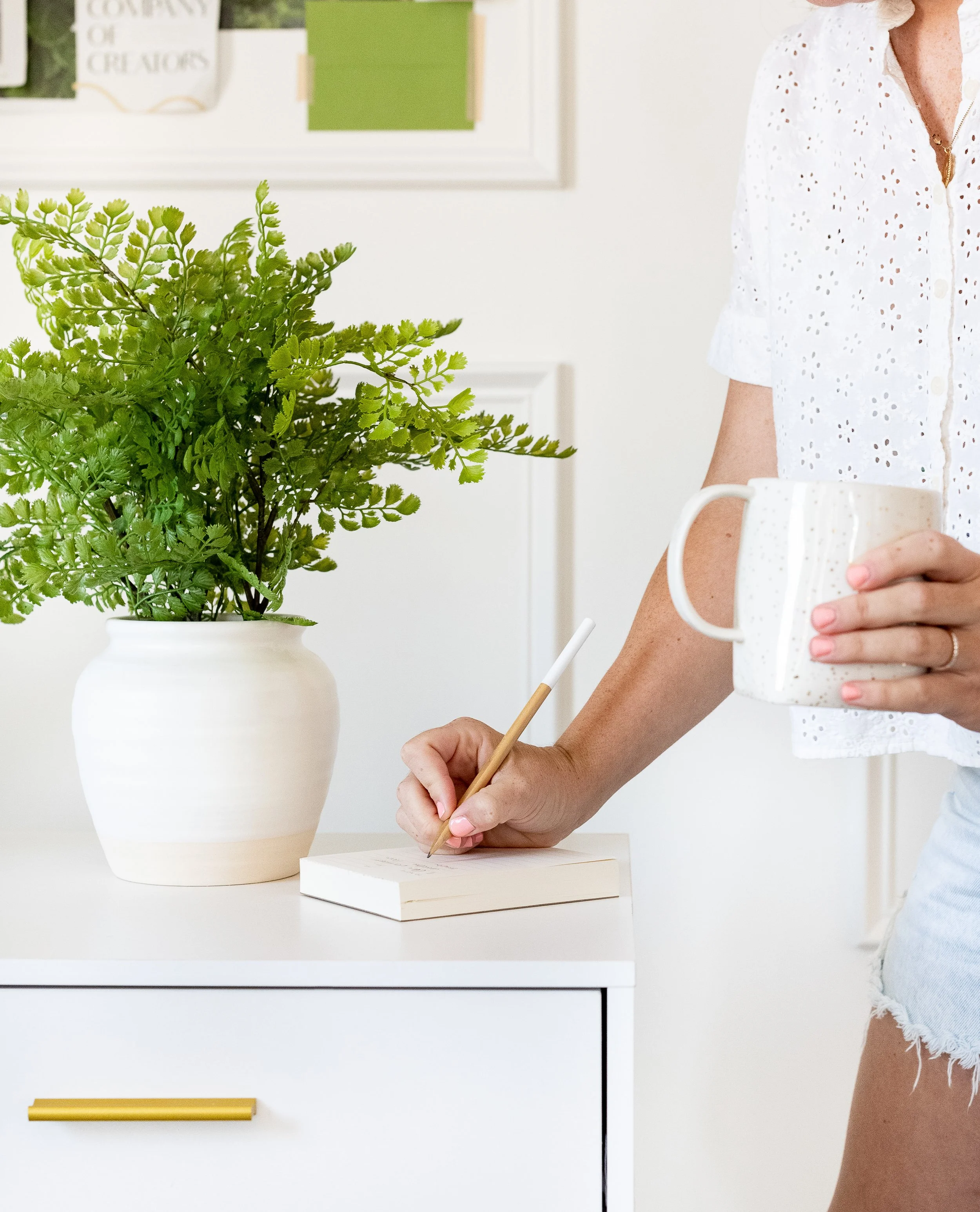 Person writing in a notebook with a mug in hand, standing next to a white dresser with a large green plant in a white pot.