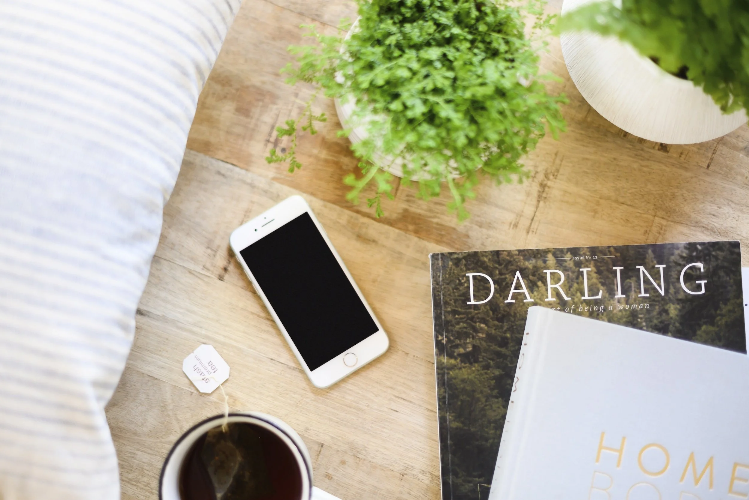 A wooden table with a potted green plant, an iPhone, a cup of tea, and magazines titled "Darling" and "Home". Part of a bed with striped bedding is visible on the left.
