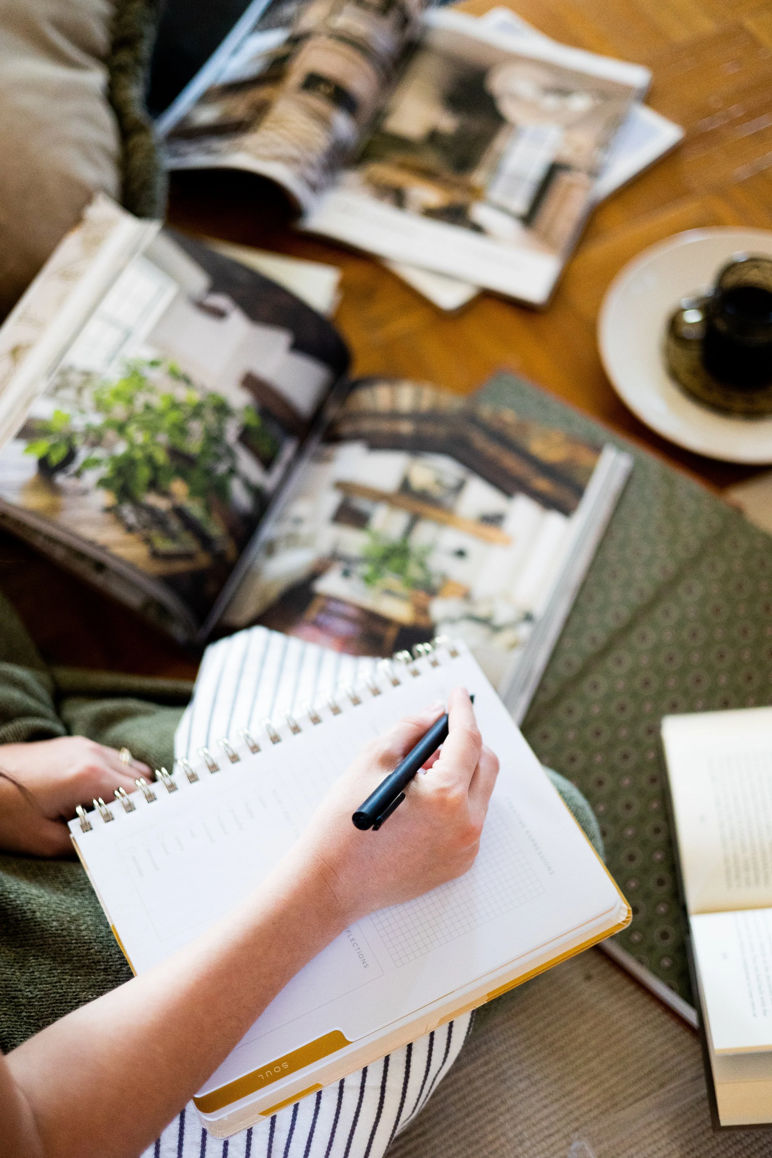 Person taking notes on a spiral notebook while surrounded by open magazines and books on a wooden table, with a cup of coffee nearby.