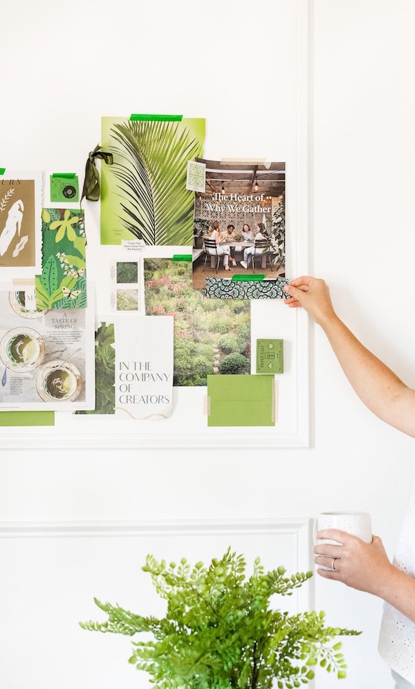 A white wall with an organized corkboard display of magazines, photos, and green swatches, a person's hand pinning a magazine, and another hand holding a white mug, with a green potted plant in front.
