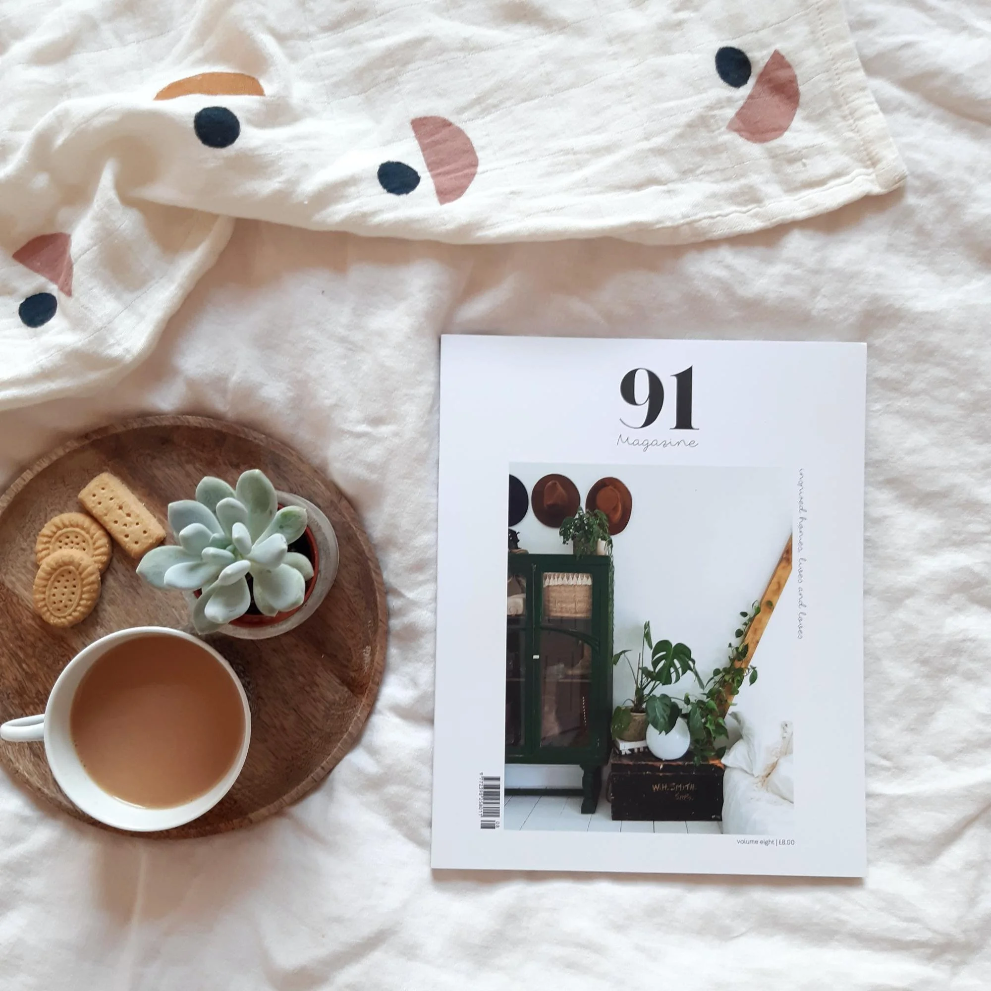 A magazine titled '91 Magazine' lying on a bed next to a wooden tray with cookies, a small succulent plant, and a cup of tea or coffee, with a white blanket with painted face designs in the background.