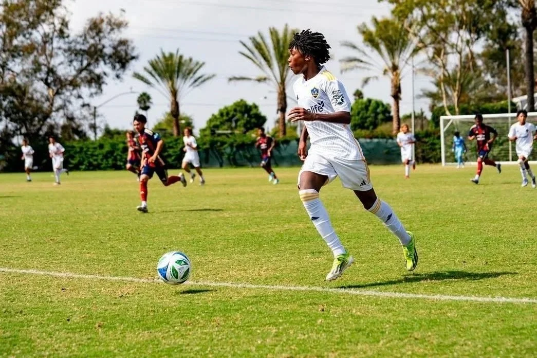 Soccer player in a white uniform running with a soccer ball on a grassy field, with other players and goalpost in the background.