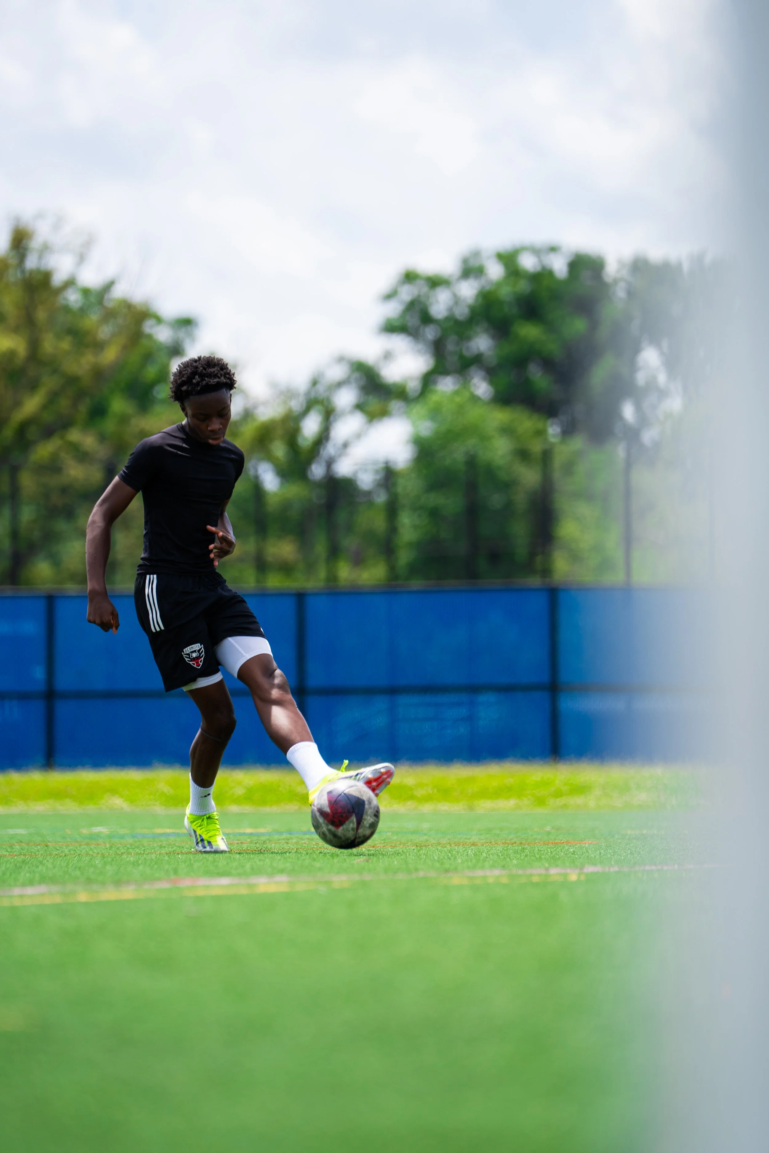 A young male soccer player in black sports attire practicing on a green field, kicking a soccer ball.