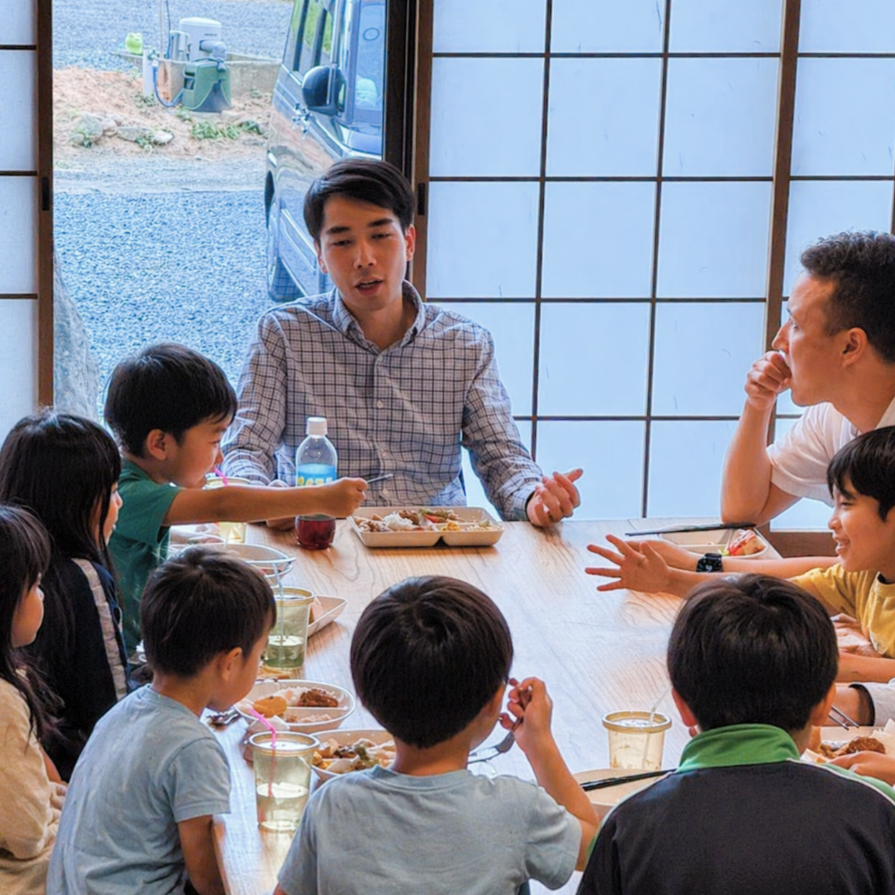 子供たちと大人のグループが和風の部屋で食事をしている様子。窓の外に車と庭が見える。男性が話している。