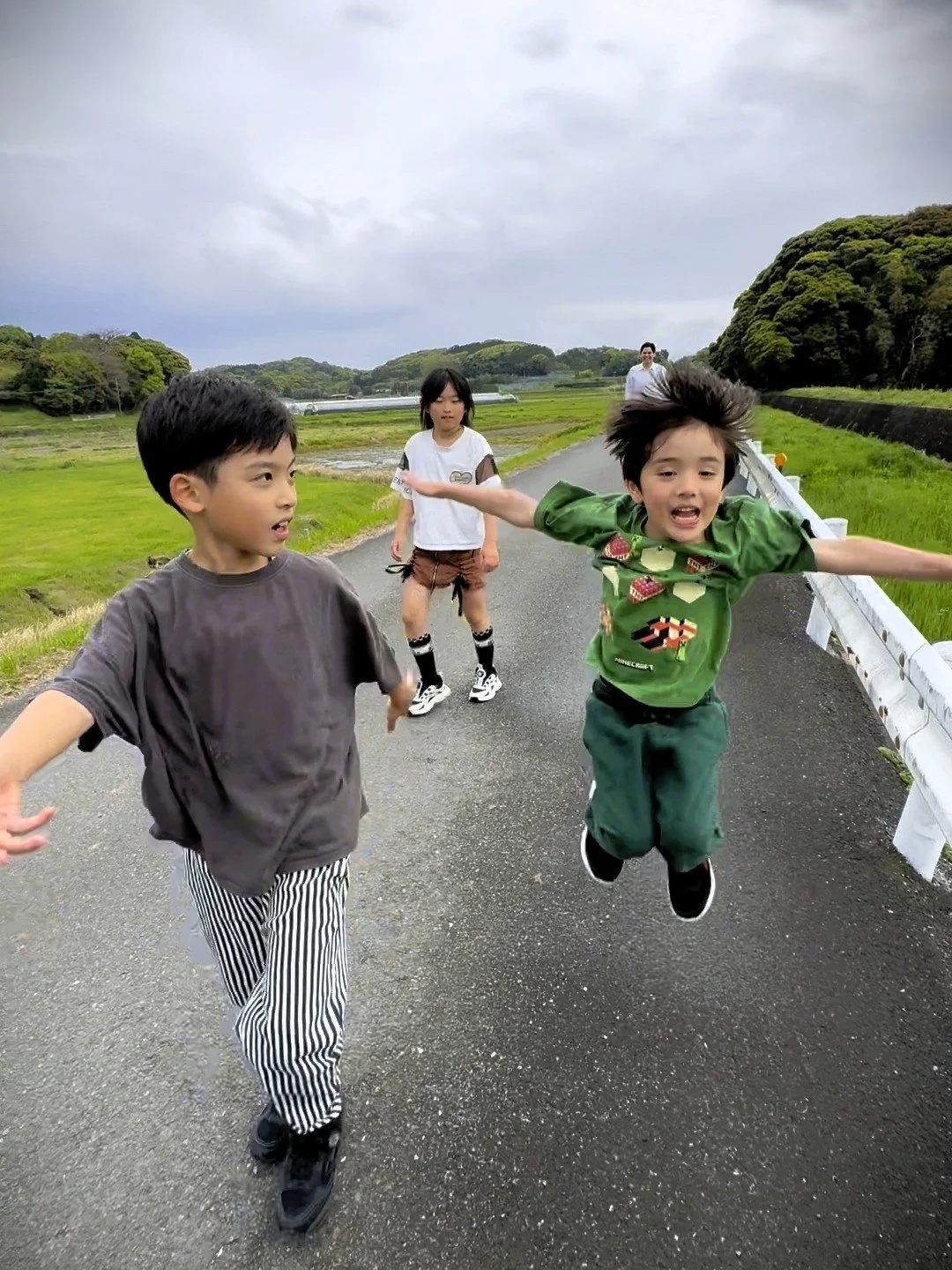 4人の子供が走っている風景。緑の田んぼの中の道路を背景に、青空と一部曇り空が広がる。