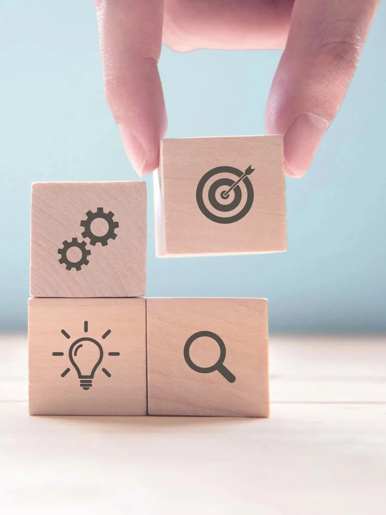 Four wooden blocks arranged in a square with icons of gears, a light bulb, a magnifying glass, and a target with an arrow.