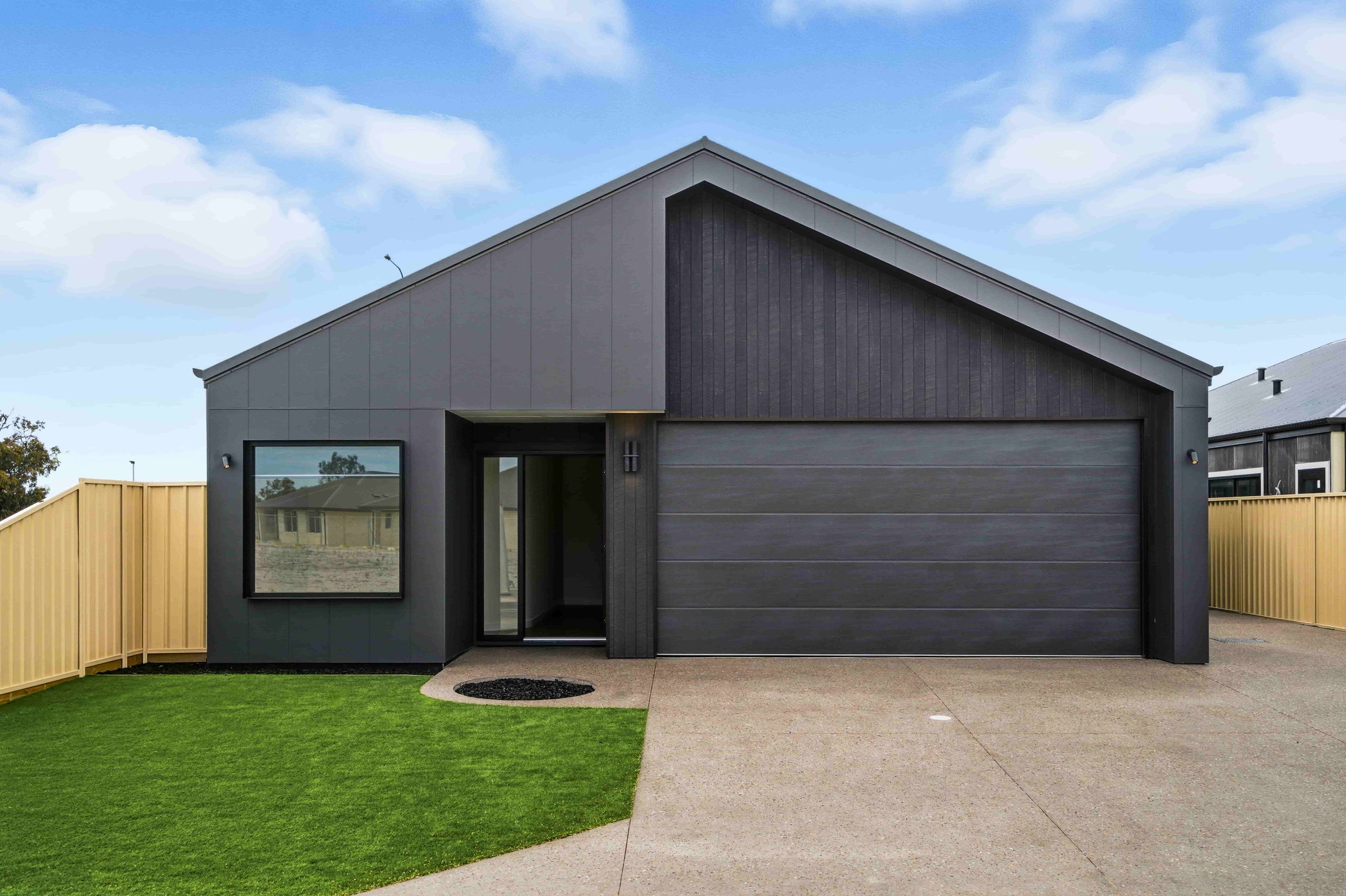 Modern house with gray exterior, large garage door, front entrance with a glass door, window reflecting neighboring houses, yellow fence on the left, well-kept green lawn, and concrete driveway under a partly cloudy sky.