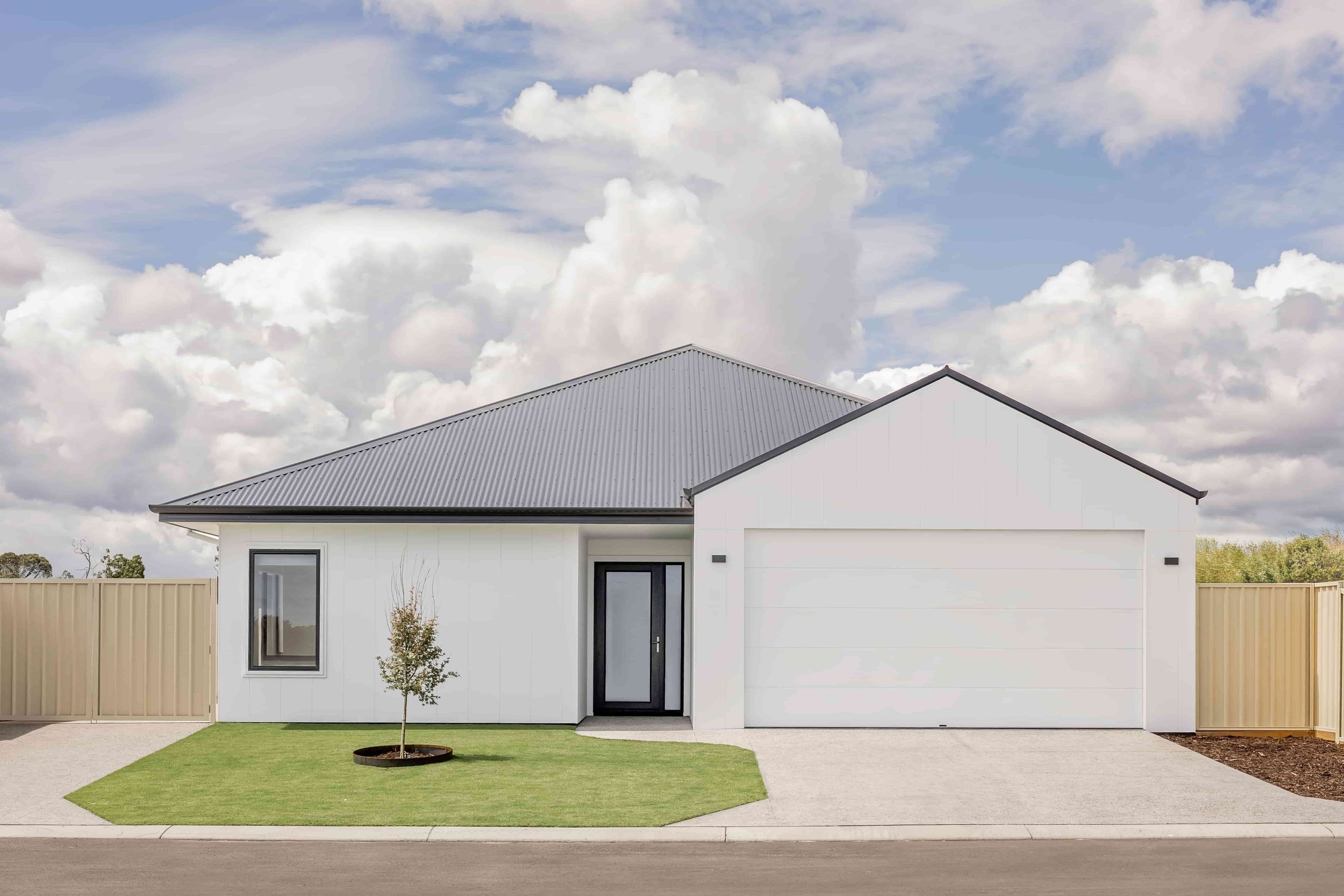 Modern white house with a gray metal roof, front door, large garage, small tree in front yard, surrounded by a beige fence, on a street with a cloudy sky.