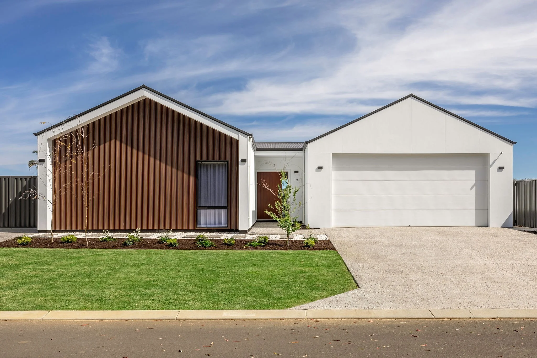 Modern single-story house with white walls, brown wood accent wall, and a two-car garage, situated in a suburban neighborhood with a green lawn and blue sky.