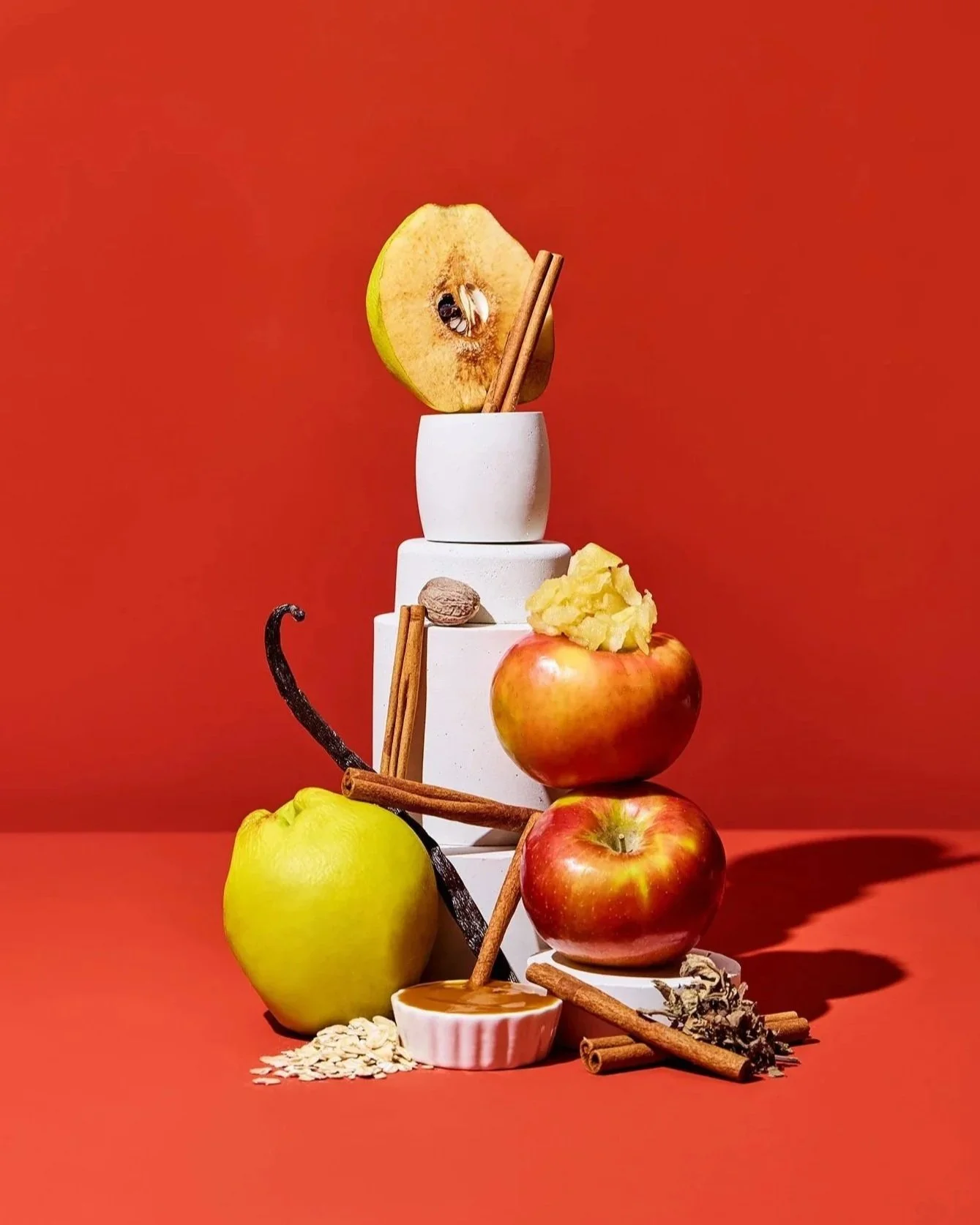 Stacked white cups with apples, cinnamon sticks, a honey dipper, and oats arranged around them against a red background.