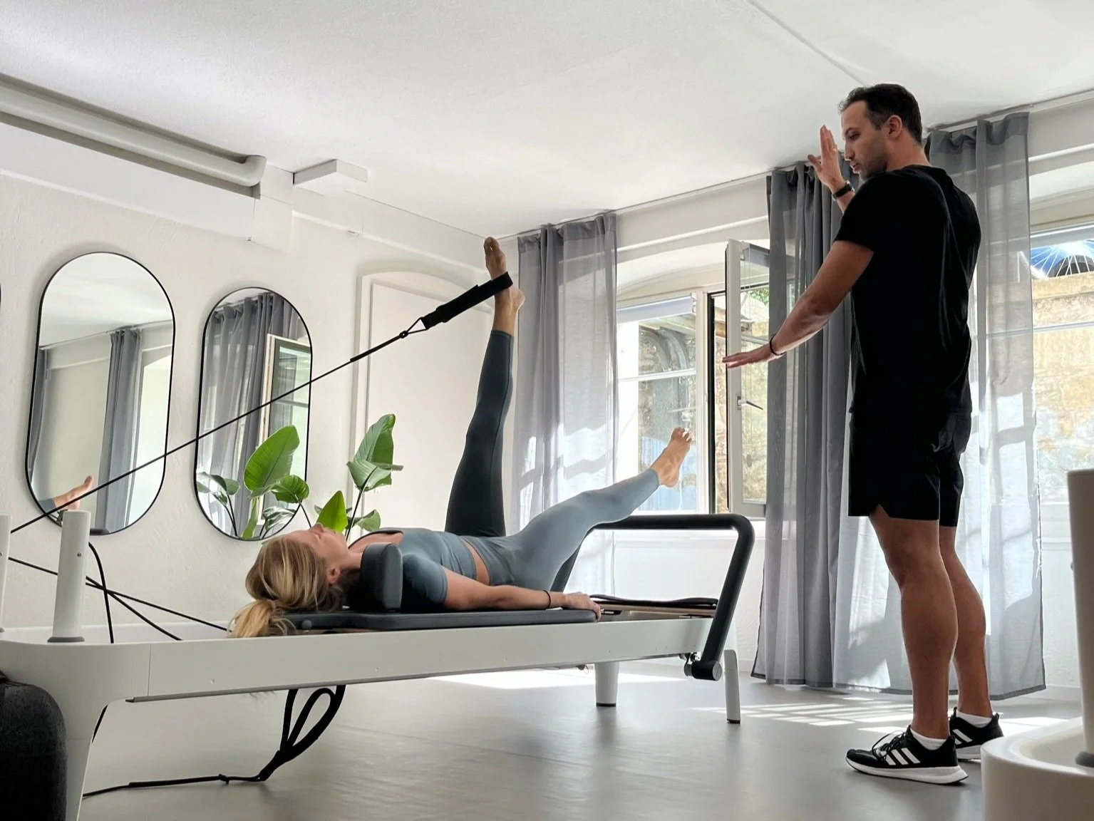 A woman performing a Pilates exercise on a reformer machine with leg stretches, supervised by a male trainer in a bright room with mirrors and windows.