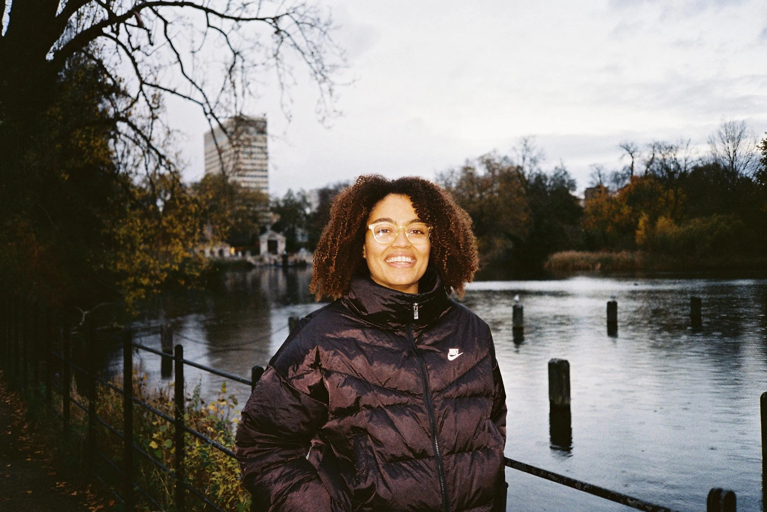 A woman with curly hair and glasses, smiling, standing by a river in a park during overcast weather, wearing a black Nike jacket. Trees with autumn foliage and a city building are visible in the background.