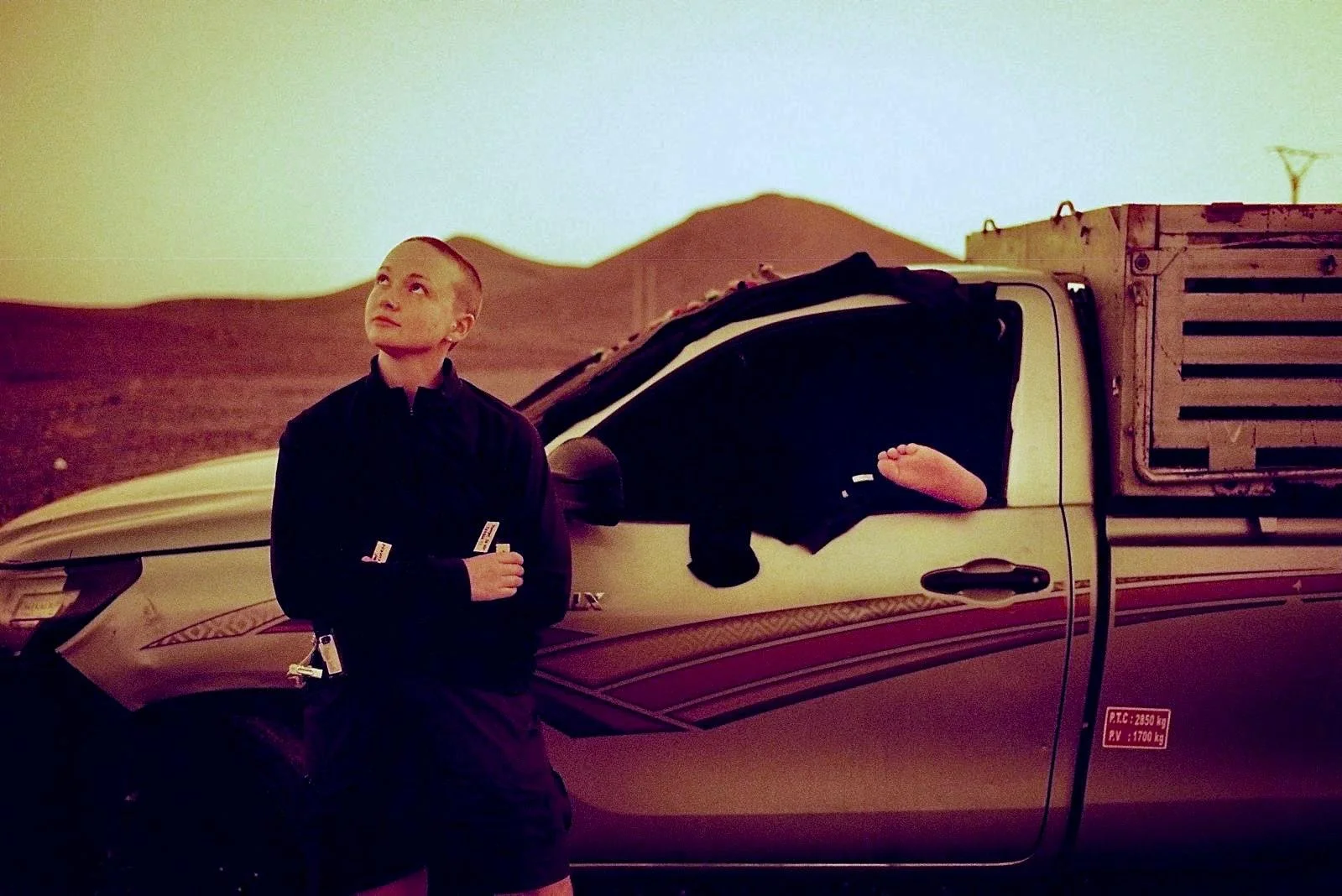 Young person standing next to a car in a desert landscape, looking up with a contemplative expression.