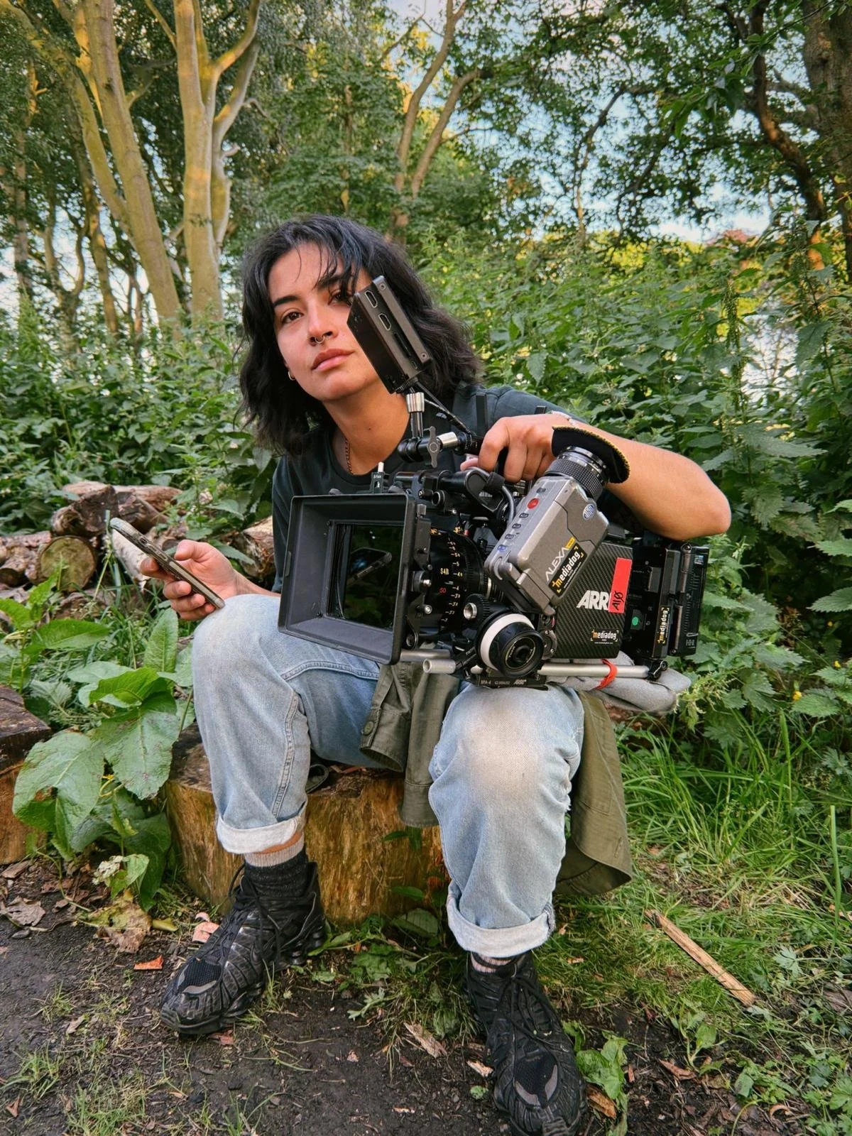 A young woman with black hair and a septum piercing sitting outdoors surrounded by green trees and plants, holding a professional film camera.