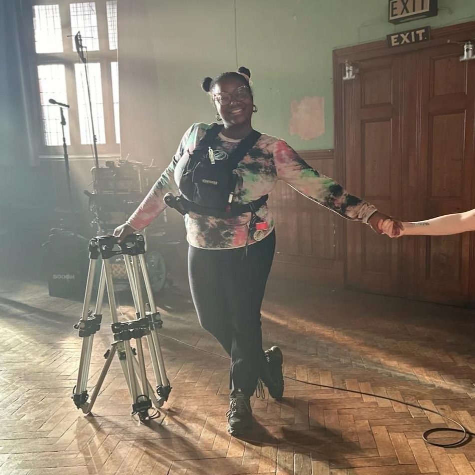 A woman standing in a room, smiling, holding hands with someone out of frame, with a tripod and microphone stand in the background, sunlight streaming through the windows.