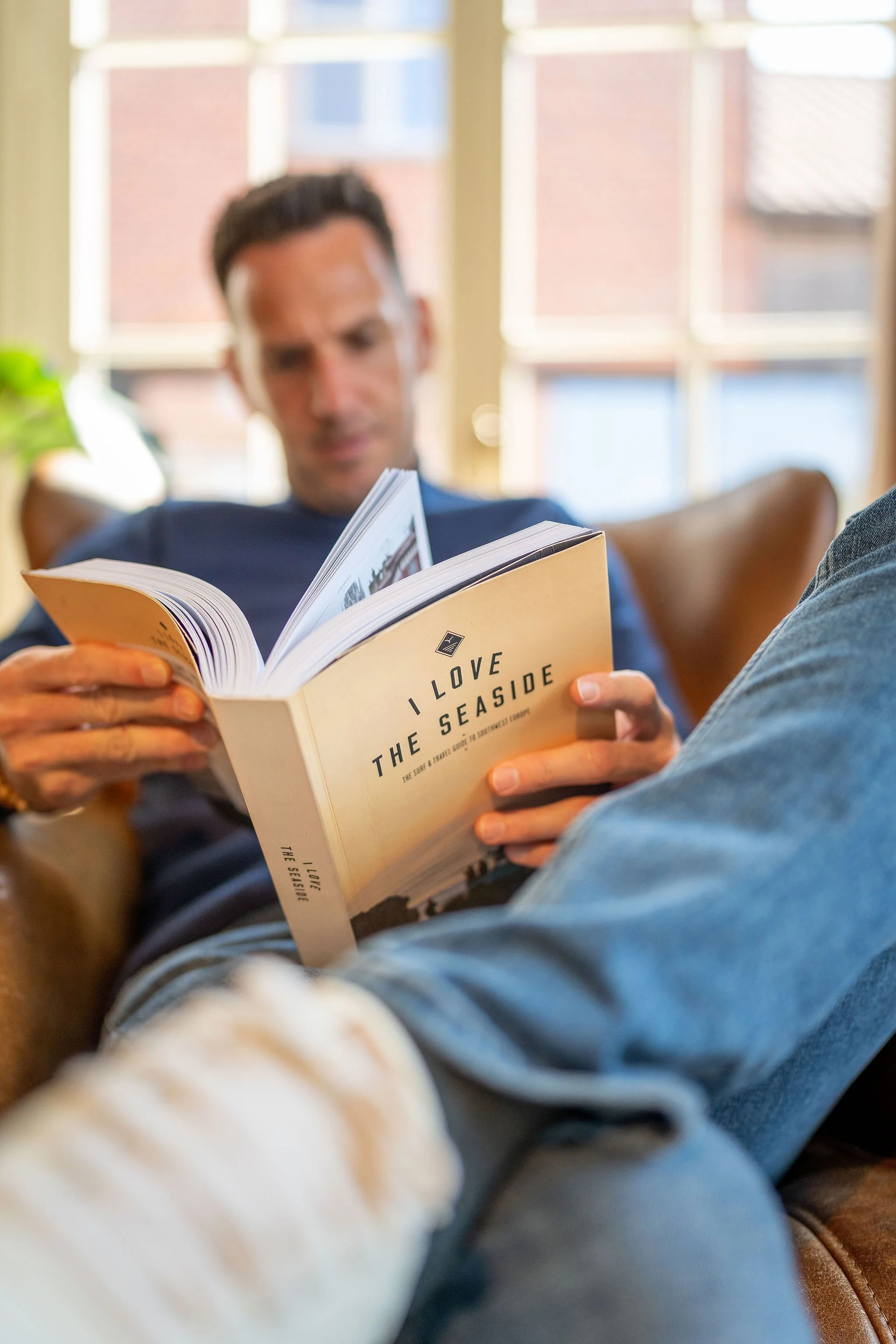 Man leest een boek met de titel 'I Love The Seaside' in een gezellige woonruimte met grote ramen.