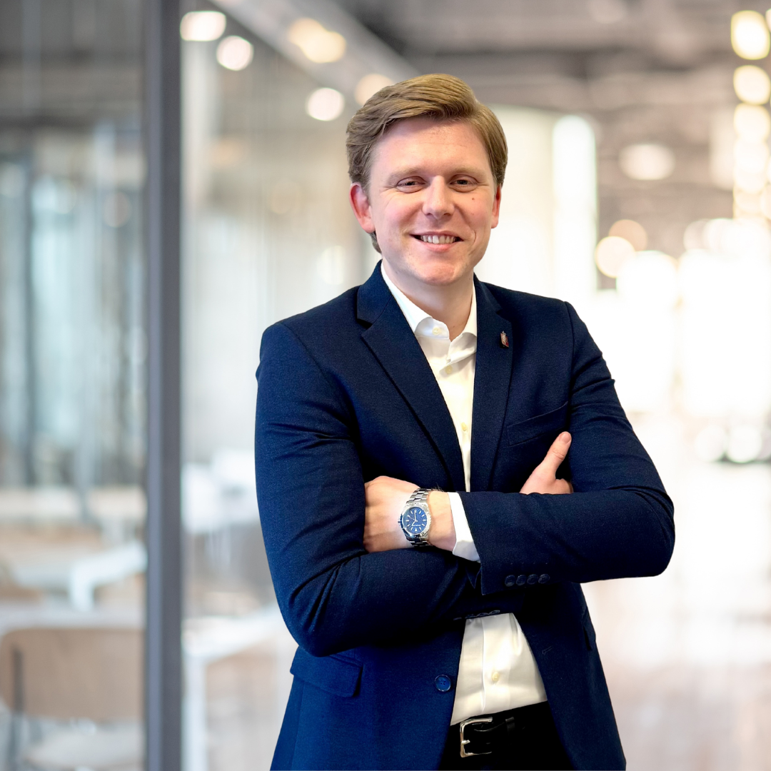 En mand i mørkeblå jakkesæt og hvid skjorte, står med armene krydset, smilende i et moderne kontormiljø.
