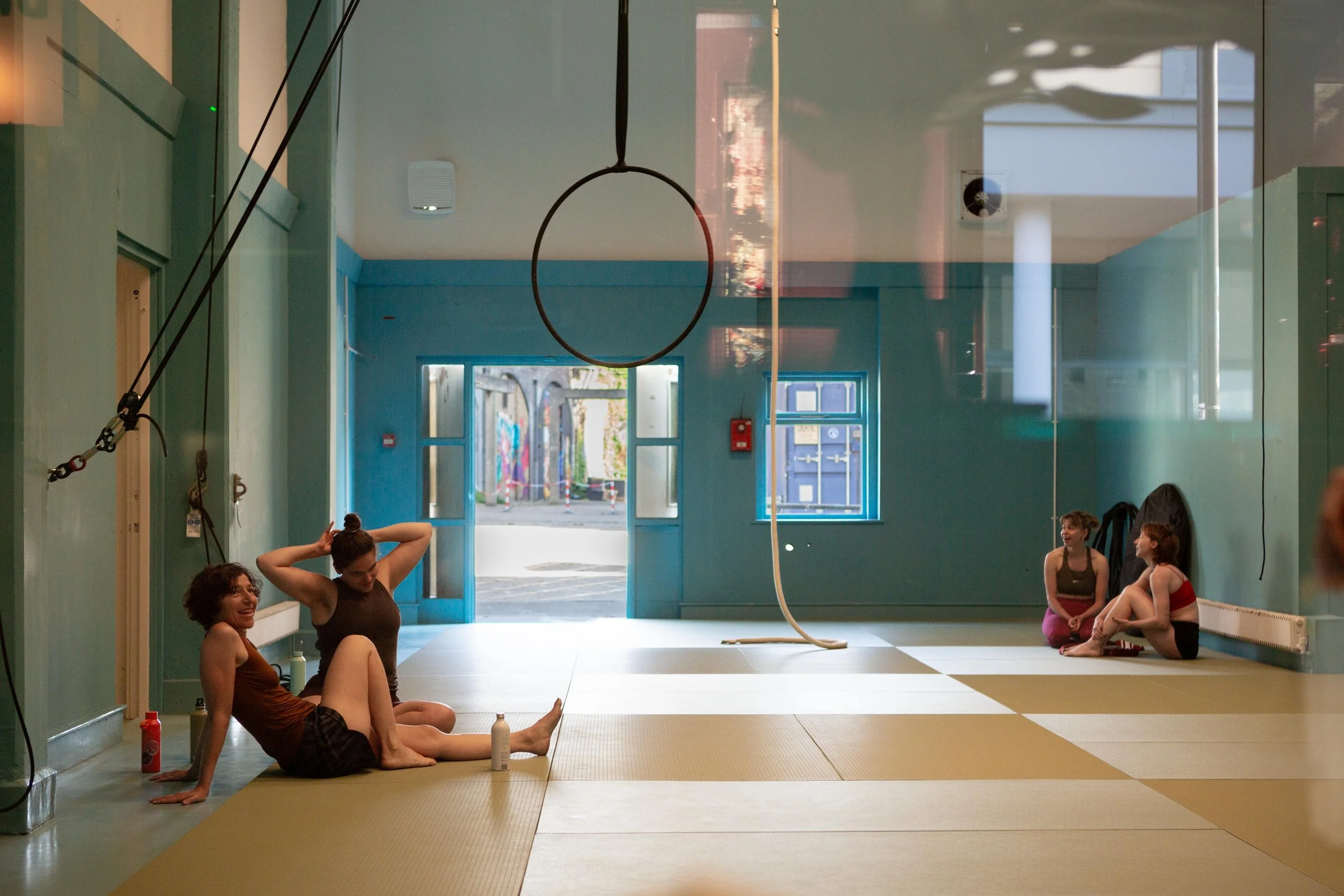 Four women sitting and talking on a tan-colored gym mat inside a light blue gym studio, with open doors and large windows showing an outdoor area.