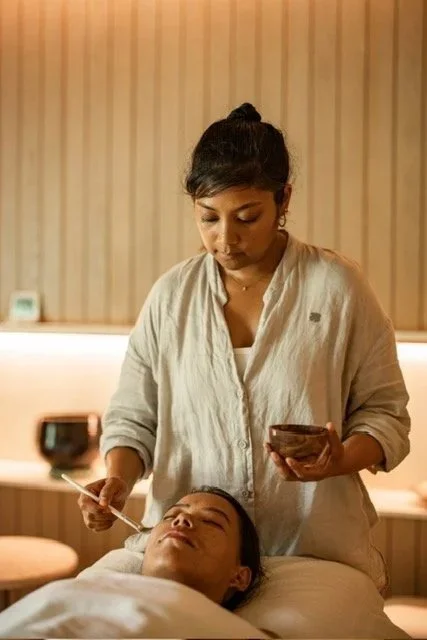 A woman lying on a treatment bed with her eyes closed, receiving a facial from a practitioner in a spa or wellness center.