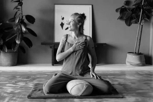 Woman practicing meditation or yoga in a room with green walls, plants, and artwork, sitting cross-legged on a mat with a bolster.
