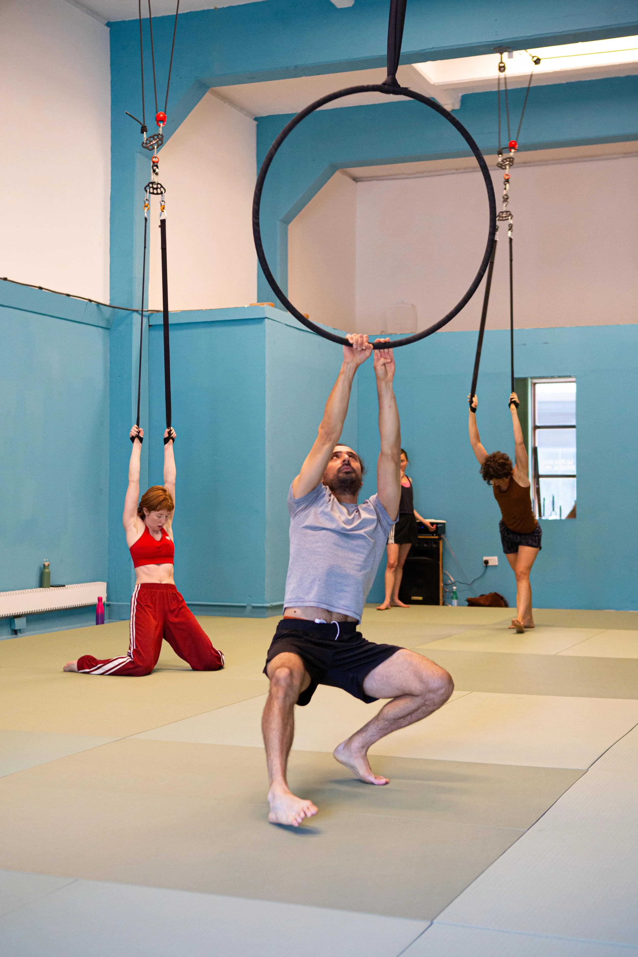 People practicing aerial fitness in a studio with blue walls and padded mats, lifting from aerial rings and hanging straps.