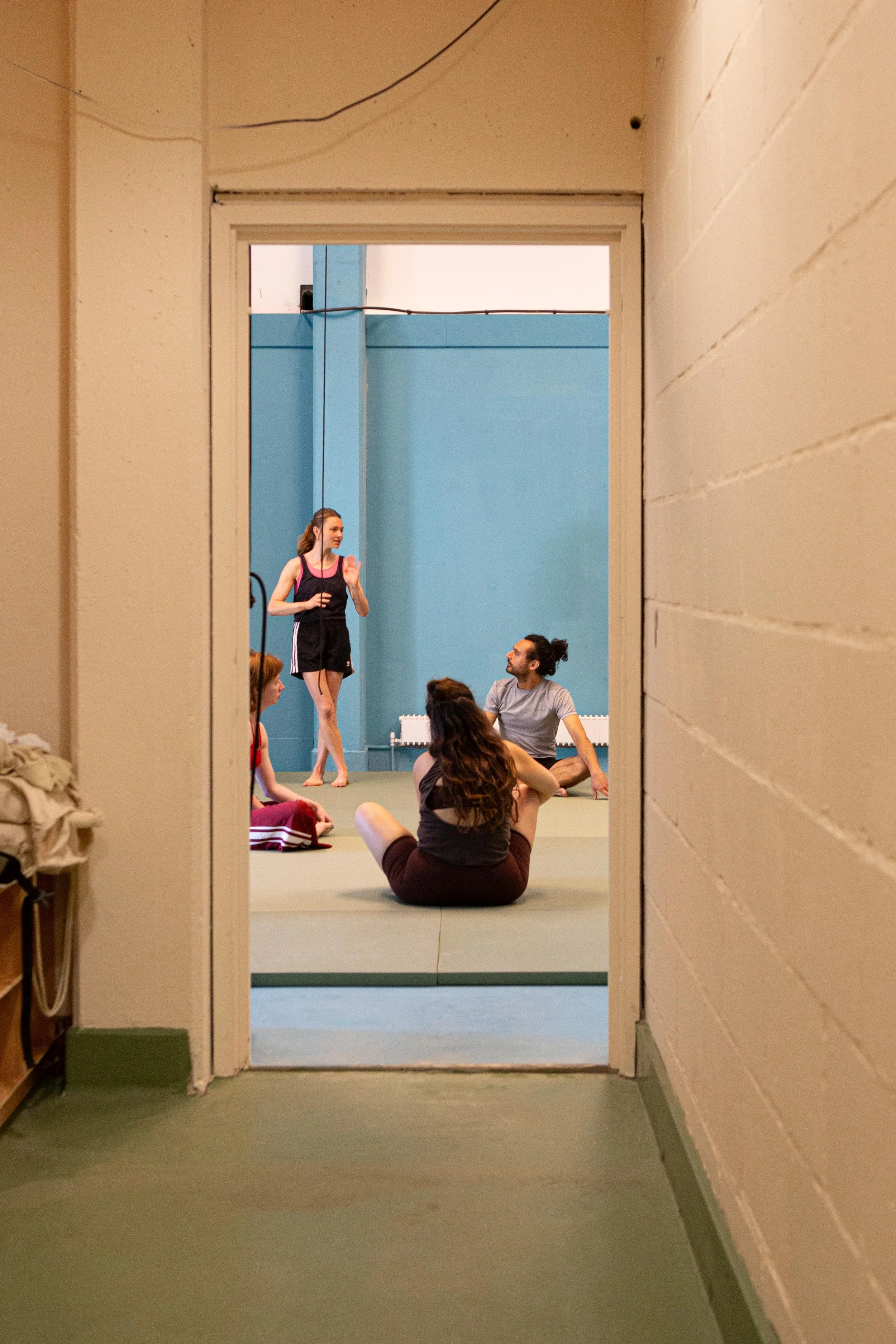 View through a doorway into a dance or exercise studio where several people are sitting on mats. One woman is standing and speaking to the group. The walls are painted blue, and there is a white radiator visible.