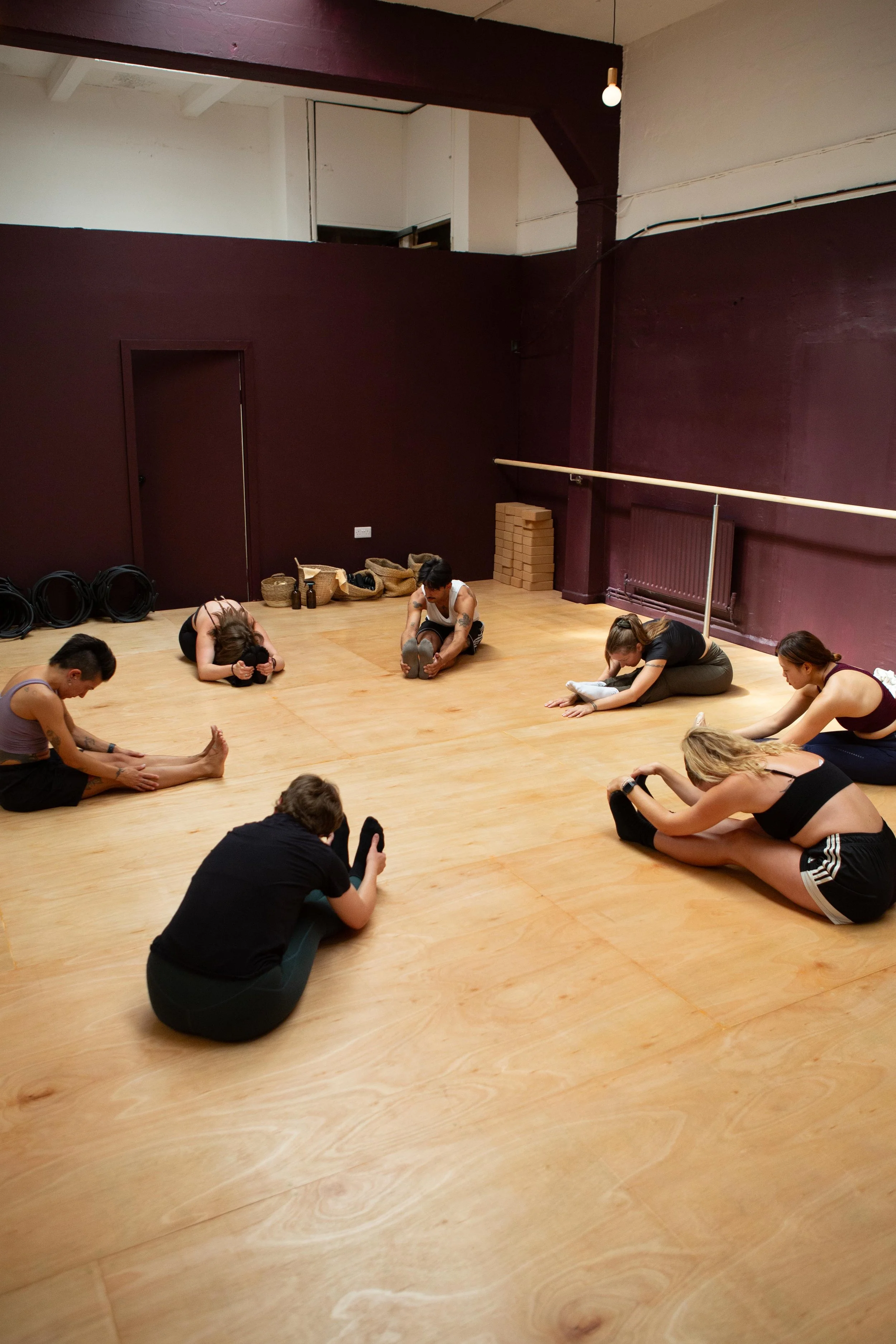 A group of six people stretching on a wooden dance studio floor, sitting with legs extended and reaching forward.
