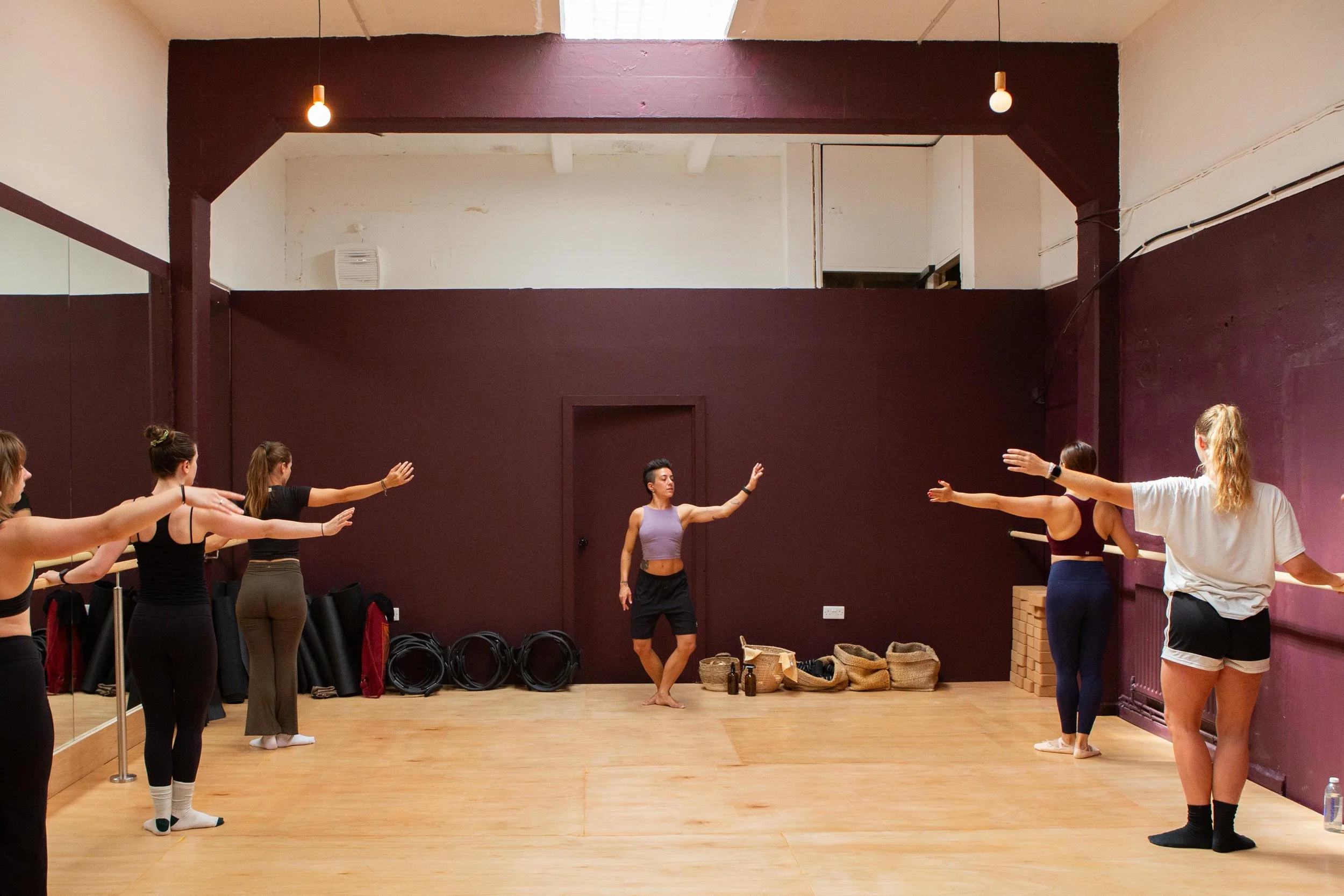 A group fitness class in a dance or yoga studio, with participants stretching and a woman instructor demonstrating movements, with ballet bars and mats visible.