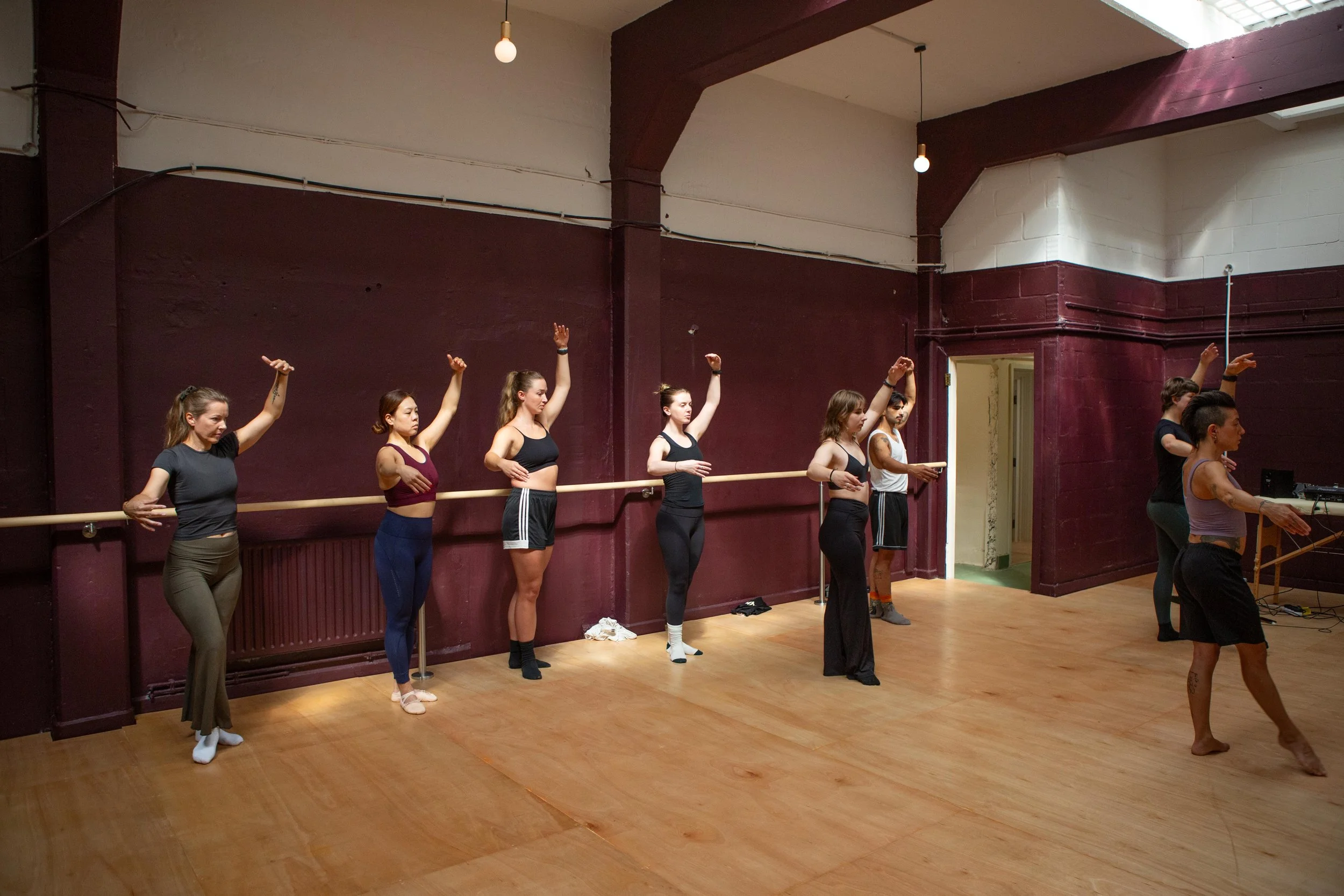 A group of women in workout clothes practicing ballet, holding a long wooden barre along the wall in a dance studio with brown walls and wooden floor.