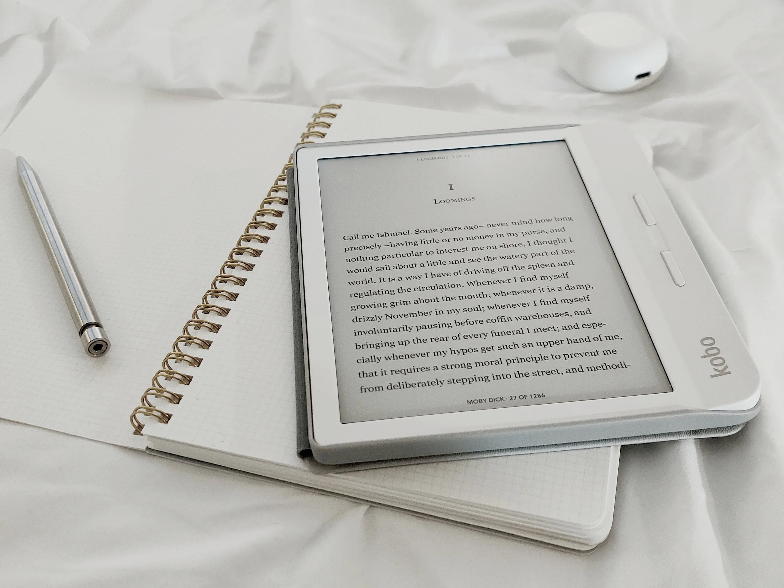 An e-reader displaying text in a white case placed on top of an open spiral notebook with white graph paper pages. To the left of the notebook, a silver pen rests on the pages. In the background, a pair of white wireless earbuds is visible on a white sheet or fabric surface.