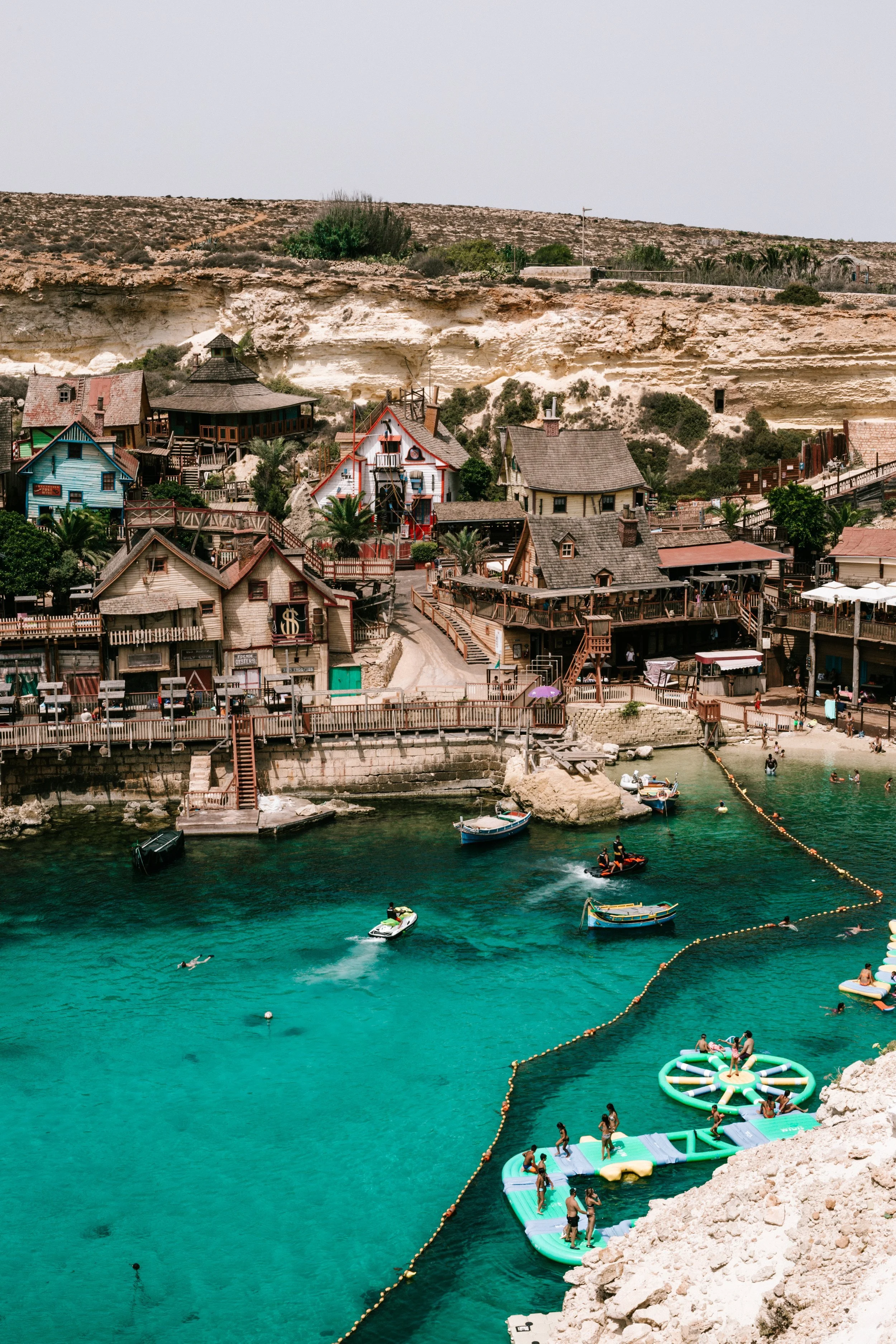 Colorful seaside village with wooden houses, boats, and people swimming and relaxing in clear turquoise water.