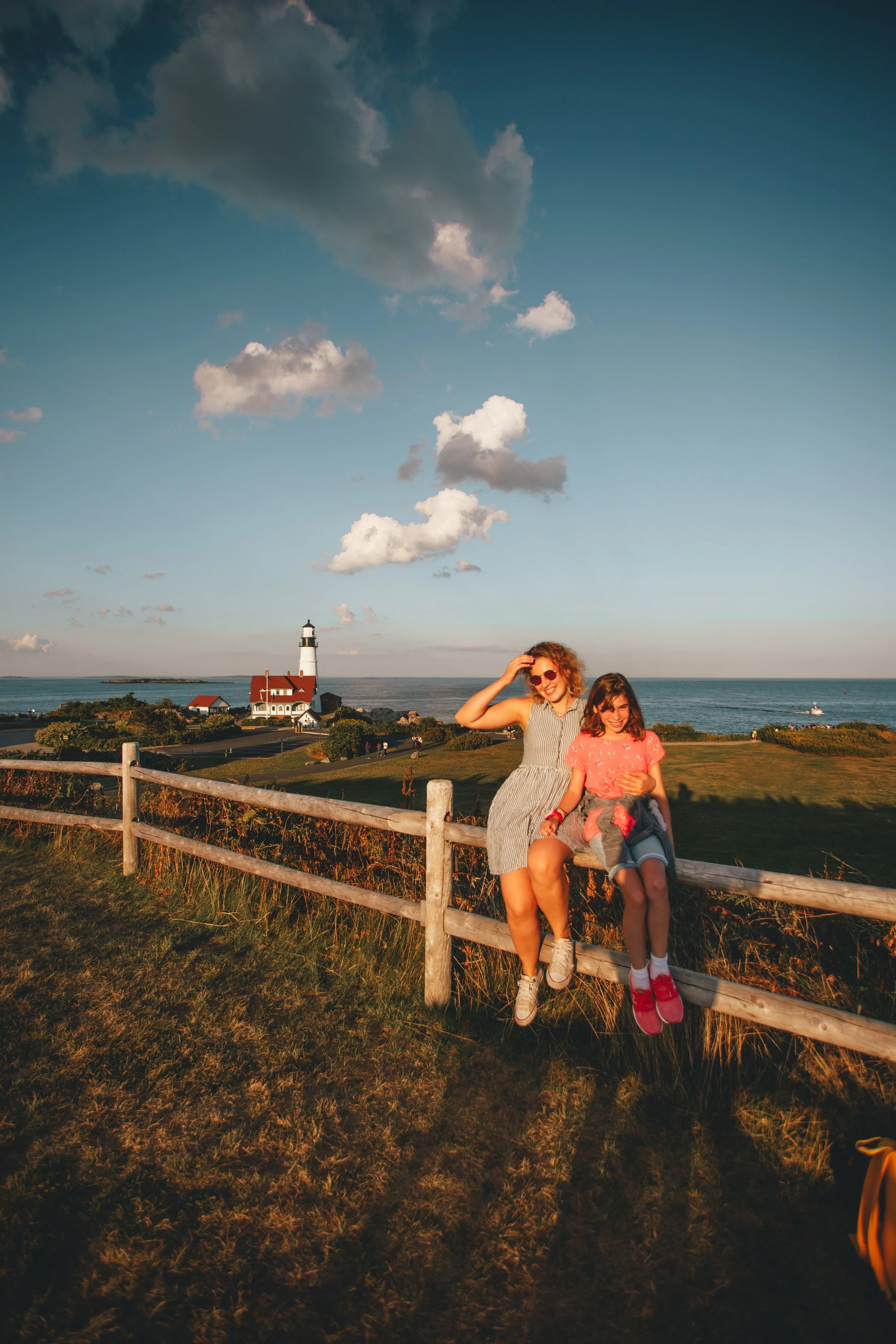 A woman and a young girl sitting on a wooden fence in front of a lighthouse by the sea during sunset.
