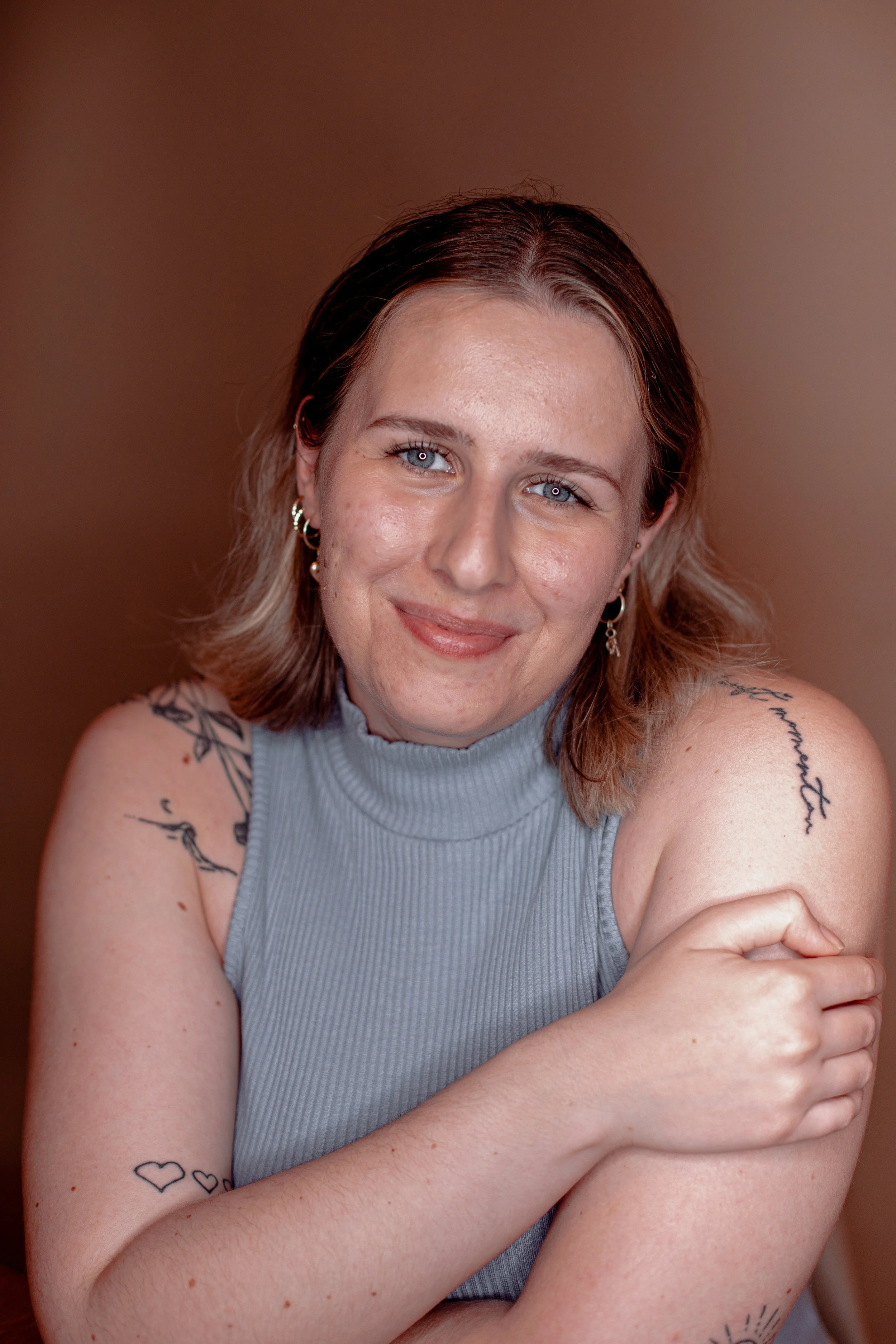 A woman with shoulder-length brown hair, blue eyes, and tattoos on her arms, wearing a sleeveless gray top, smiling at the camera.