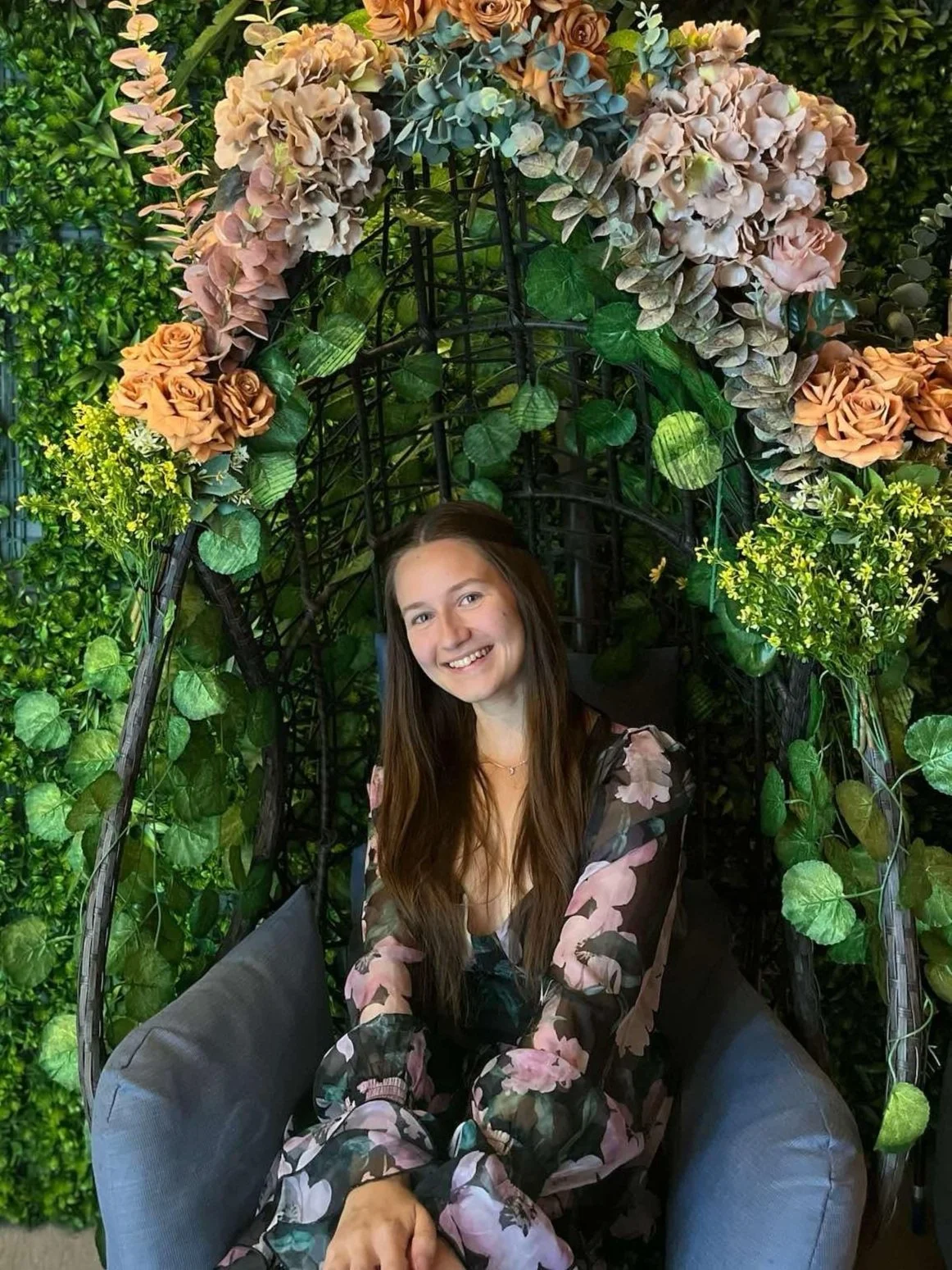 A young woman with long brown hair smiling while sitting in a decorative outdoor swing surrounded by lush green plants and topped with a floral arrangement of pink, peach, and grayish flowers.
