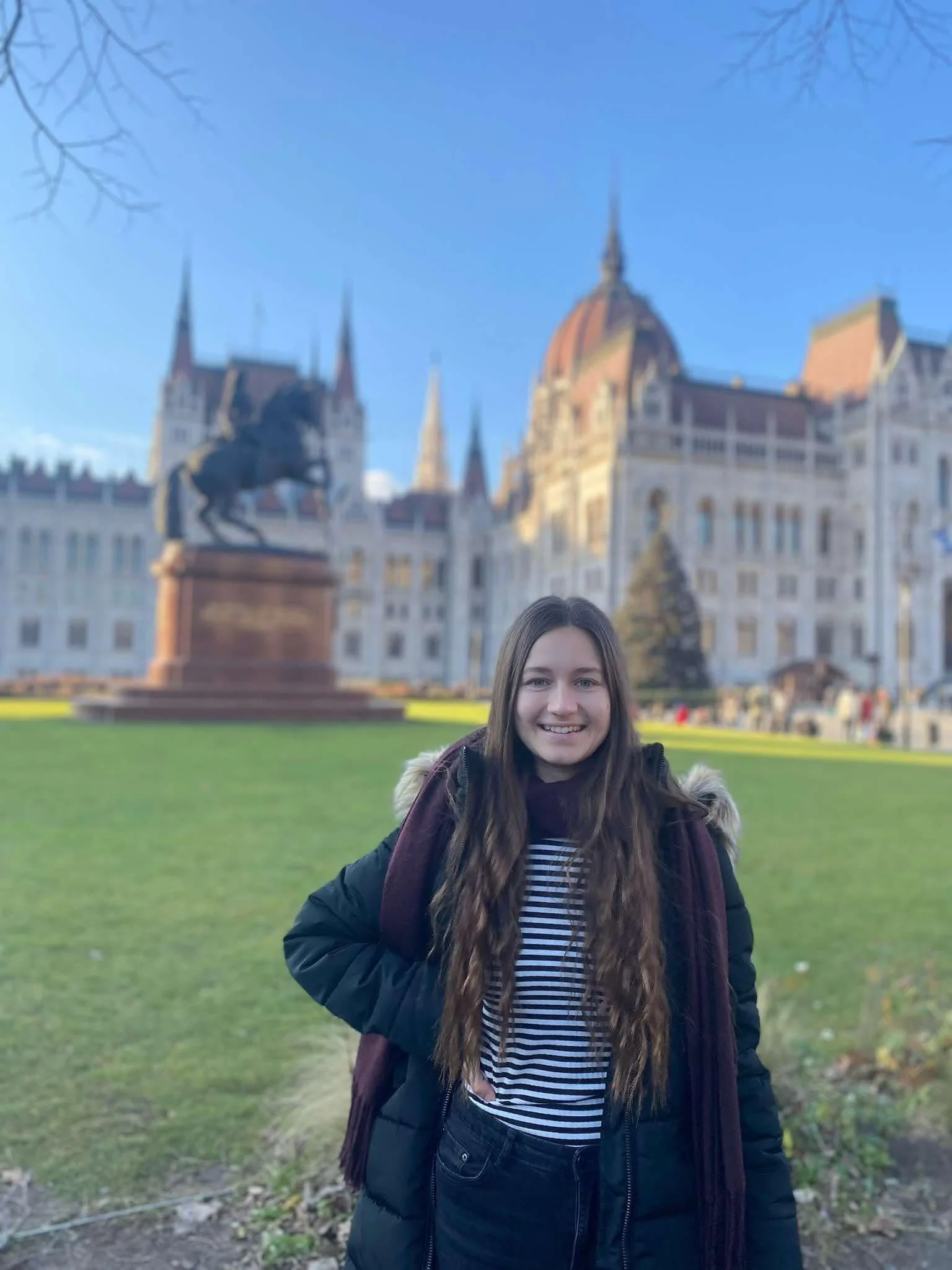 A smiling young woman standing outdoors in front of a large, historic castle with turrets and a dome, on a clear day with a blue sky.