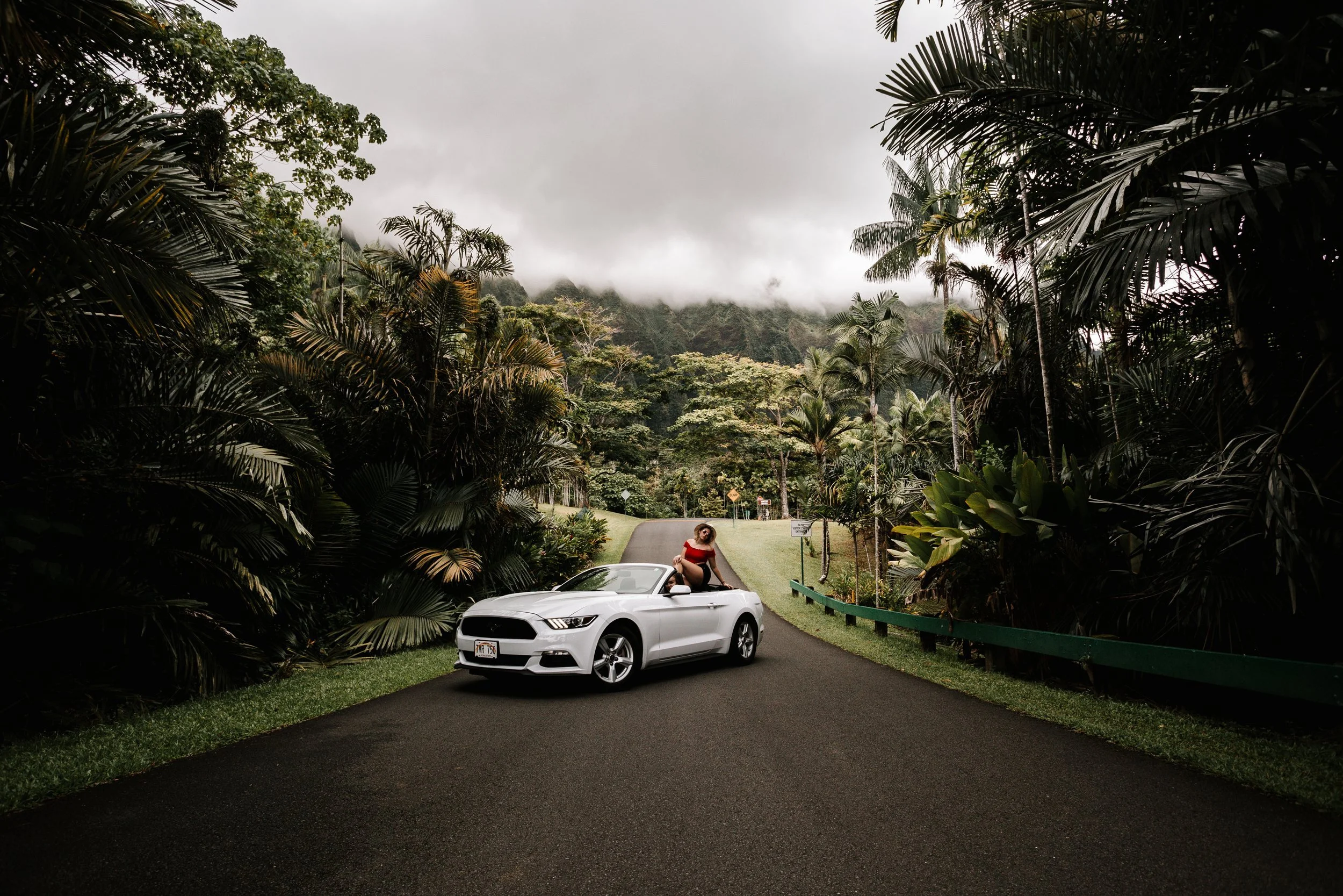 A woman in a red dress sitting on the hood of a white Ford Mustang car parked on a winding road surrounded by lush tropical vegetation, with mountains and clouds in the background.
