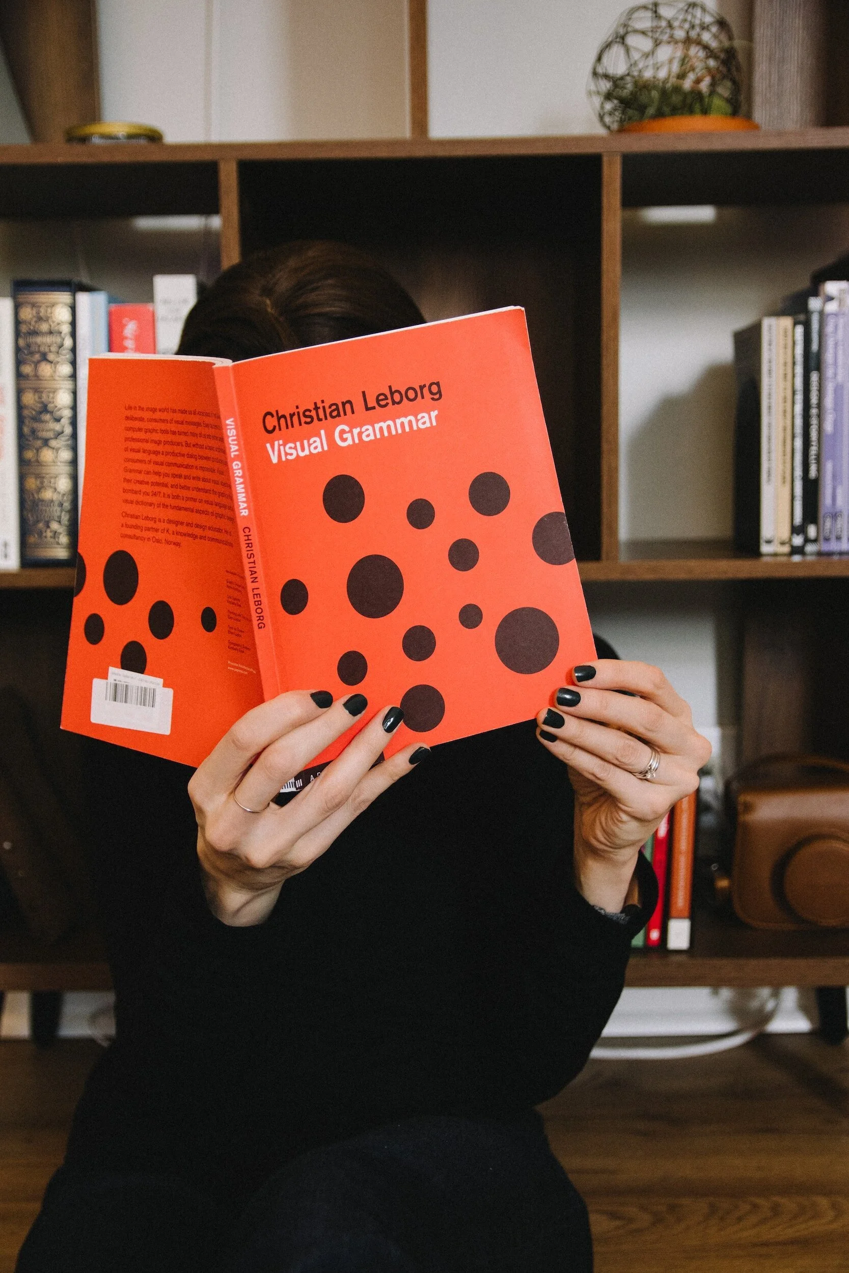 Person sitting in front of a bookshelf, holding and reading a red book titled 'Visual Grammar' by Christian Leborg, with black polka dots on the cover.