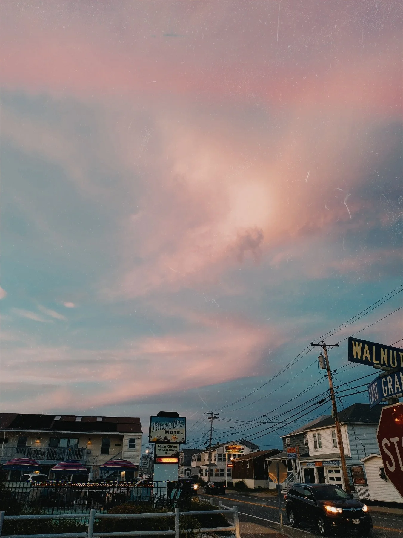 Urban street scene at dusk with pastel pink and blue sky, residential houses, a motel sign, street signs, and parked cars.