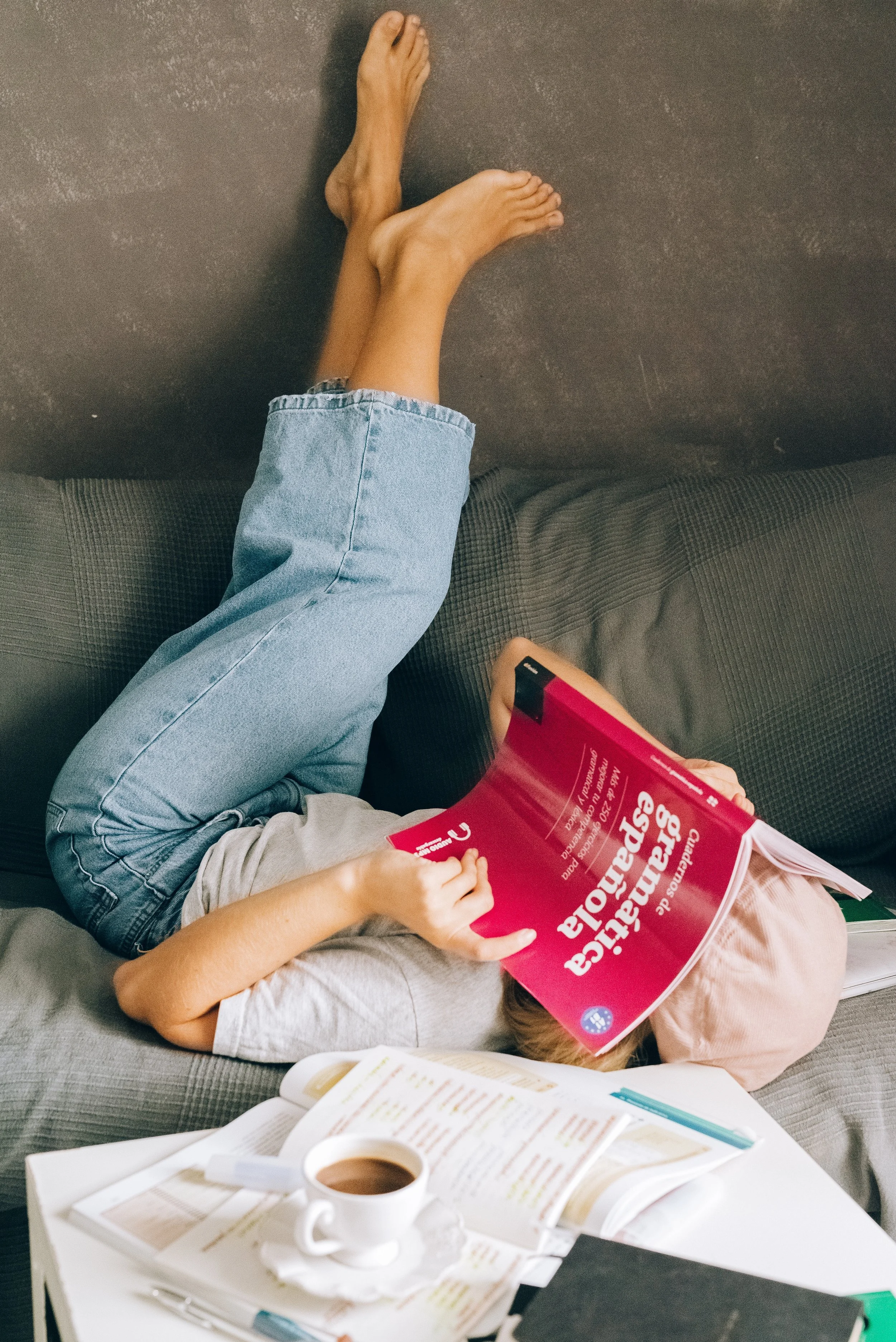 Person lying on a sofa reading a book while drinking coffee, with a notepad and pen nearby.