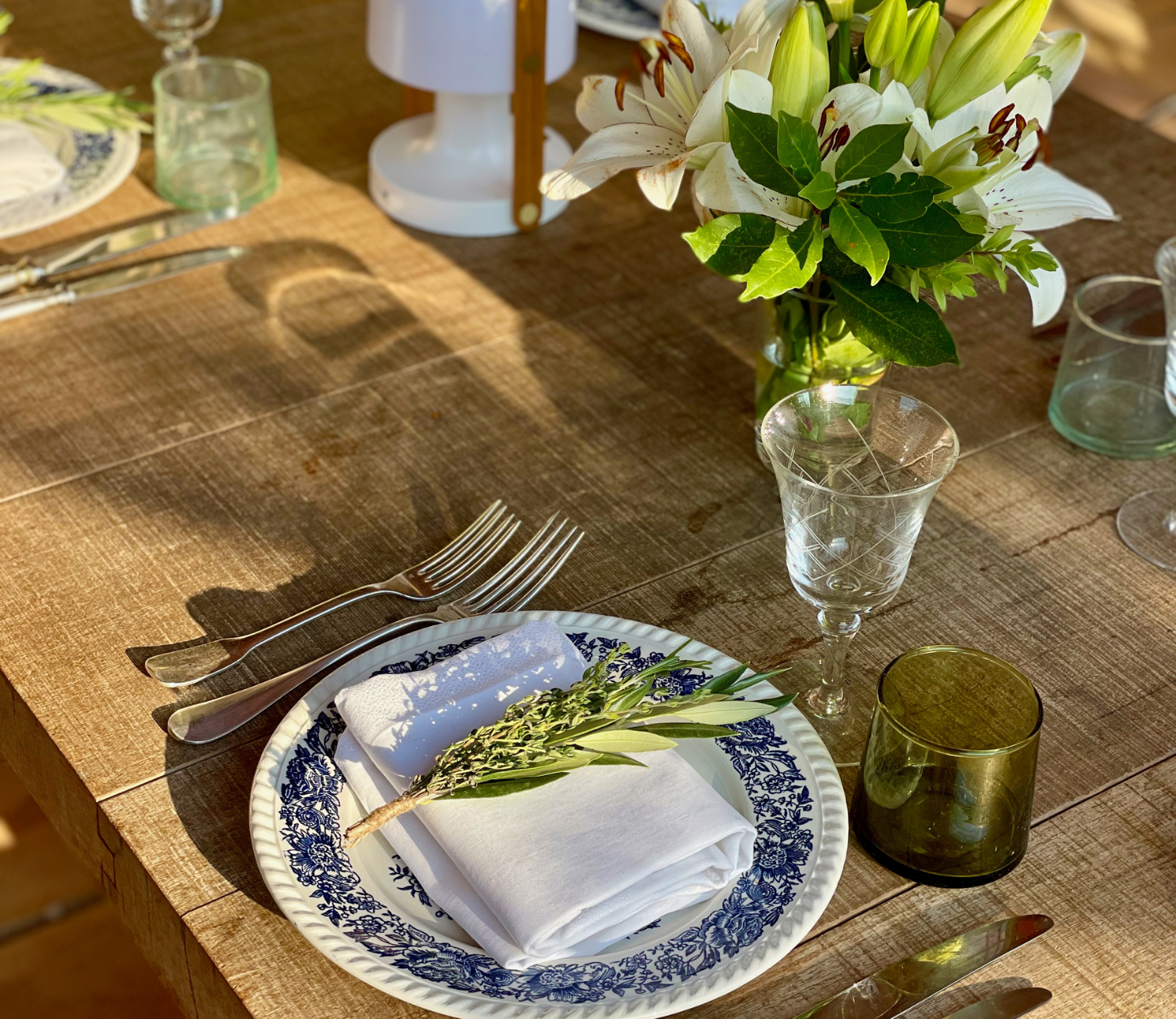 A rustic wooden dining table set with a blue and white patterned plate, a folded white napkin with a small bunch of green herbs on top, silverware, and glassware. A jar with white lilies and greenery serves as a centerpiece.