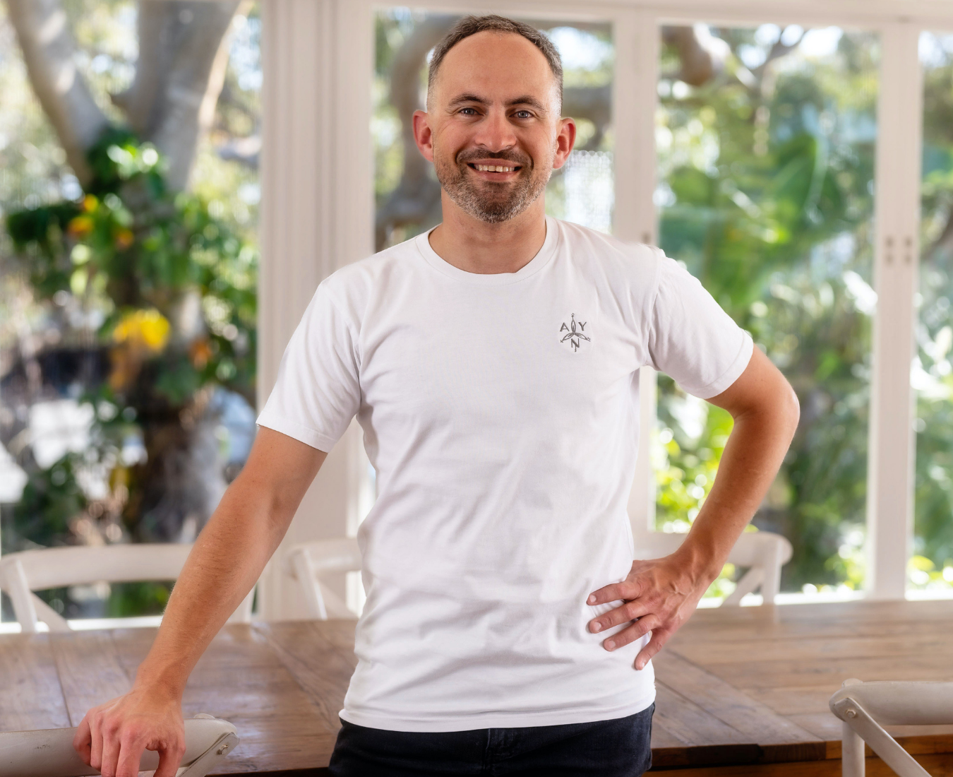Pierre Gaudin, Head chef and founder of Noranise Dining posing indoors near a wooden table, wearing a white T-shirt with a small logo on the chest, with large windows and greenery in the background.