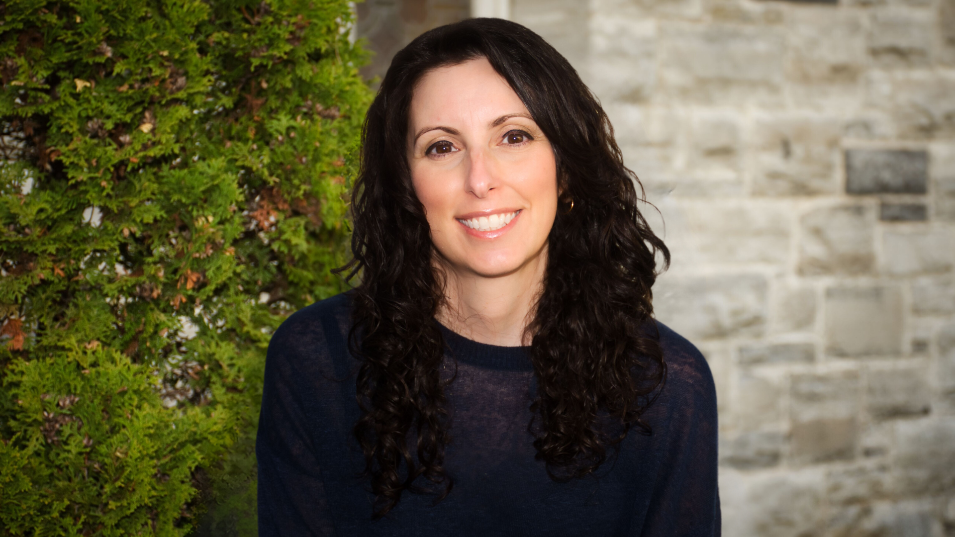 Inês Lincho, a Registered Social Worker, with long, curly dark hair smiling at the camera, standing outdoors with green shrubbery and a stone wall in the background.